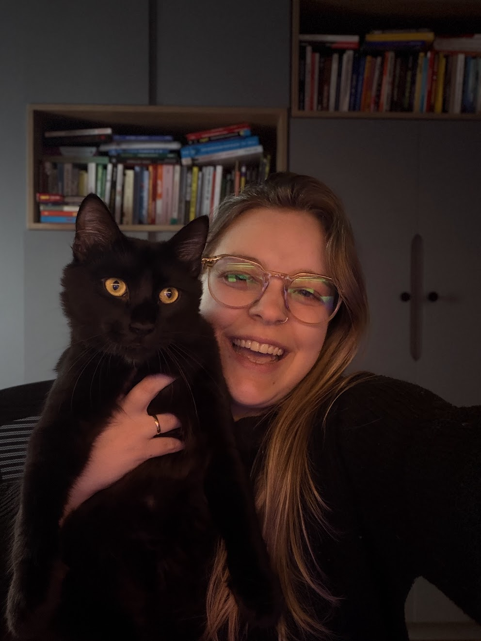 A smiling woman with glasses holding a black cat with yellow eyes, with bookshelves in the background.