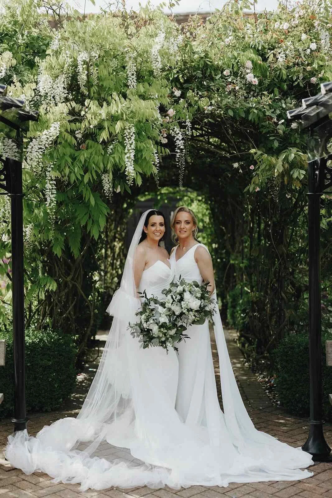 Two brides beneath the flowering wisteria in The Old Kent Barn gardens