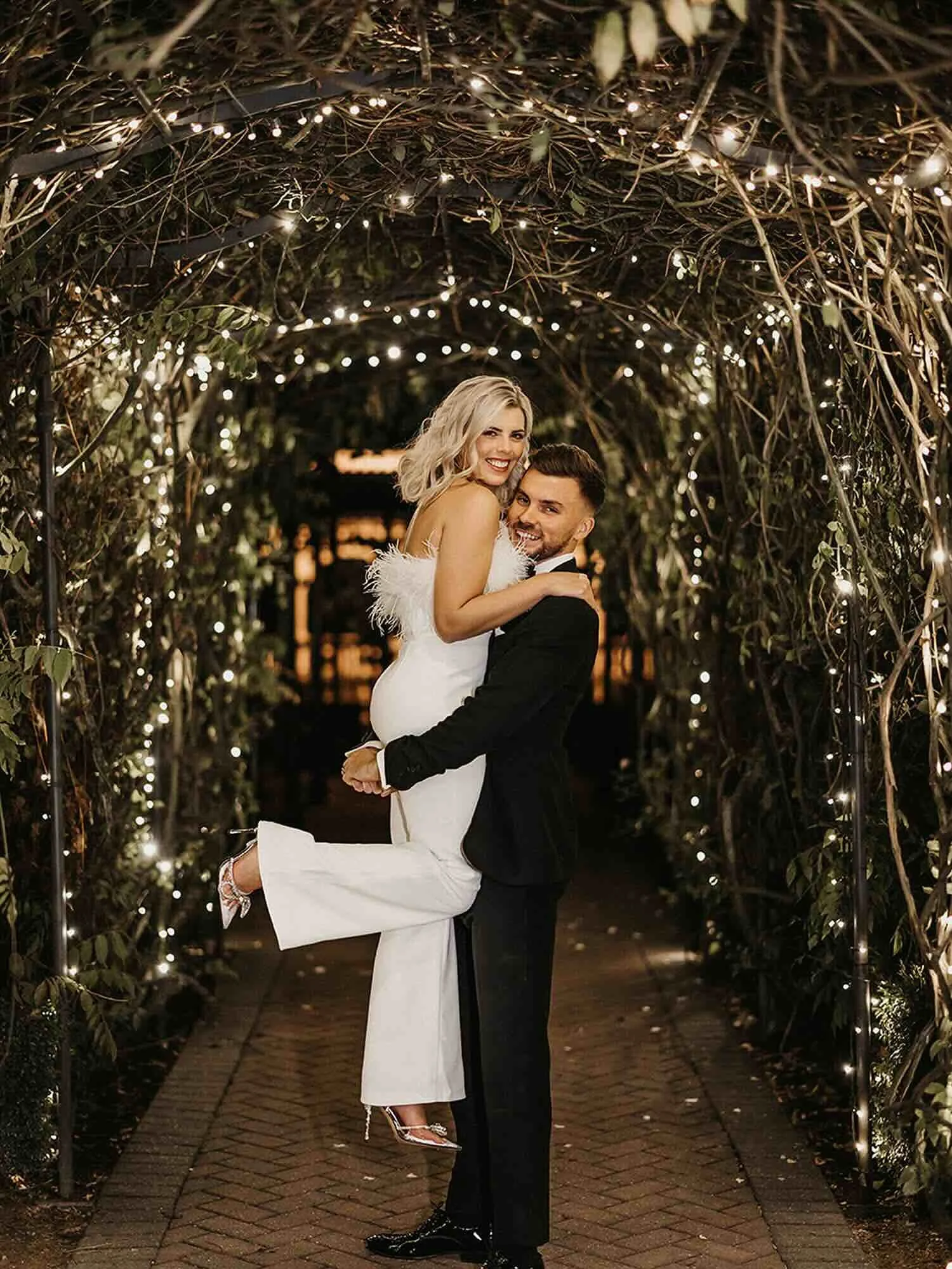 A happy couple at their wedding, with the groom lifting the bride in his arms under a decorative arch of twinkling lights at night.