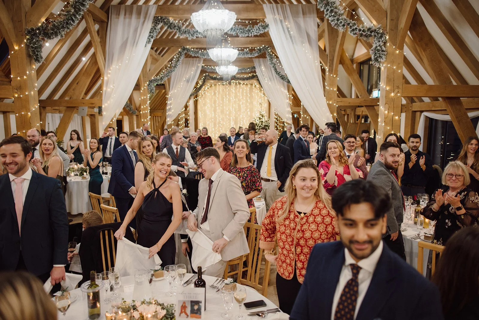 Guests at a wedding reception in a decorated wooden hall with string lights, curtains, and chandeliers, socializing and celebrating.