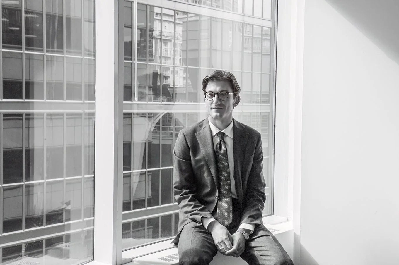 A man in business attire sitting on a windowsill in a high-rise office, with city buildings visible through the window.