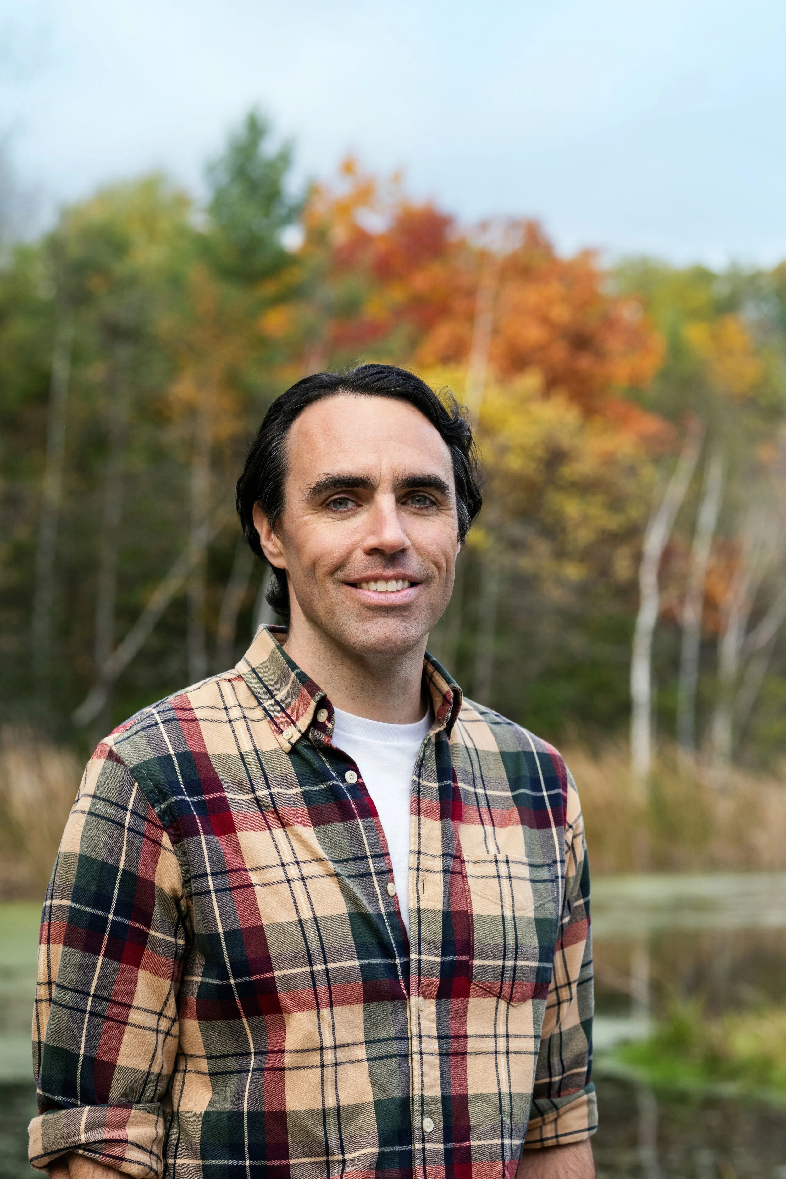 A man with dark hair and blue eyes smiling outdoors during autumn, wearing a plaid shirt with shades of beige, red, green, and blue, with a blurred background of colorful fall trees.