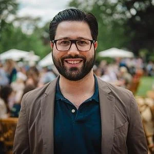 A smiling man with dark hair and glasses, wearing a brown jacket and navy shirt, standing outdoors at a park or festival.