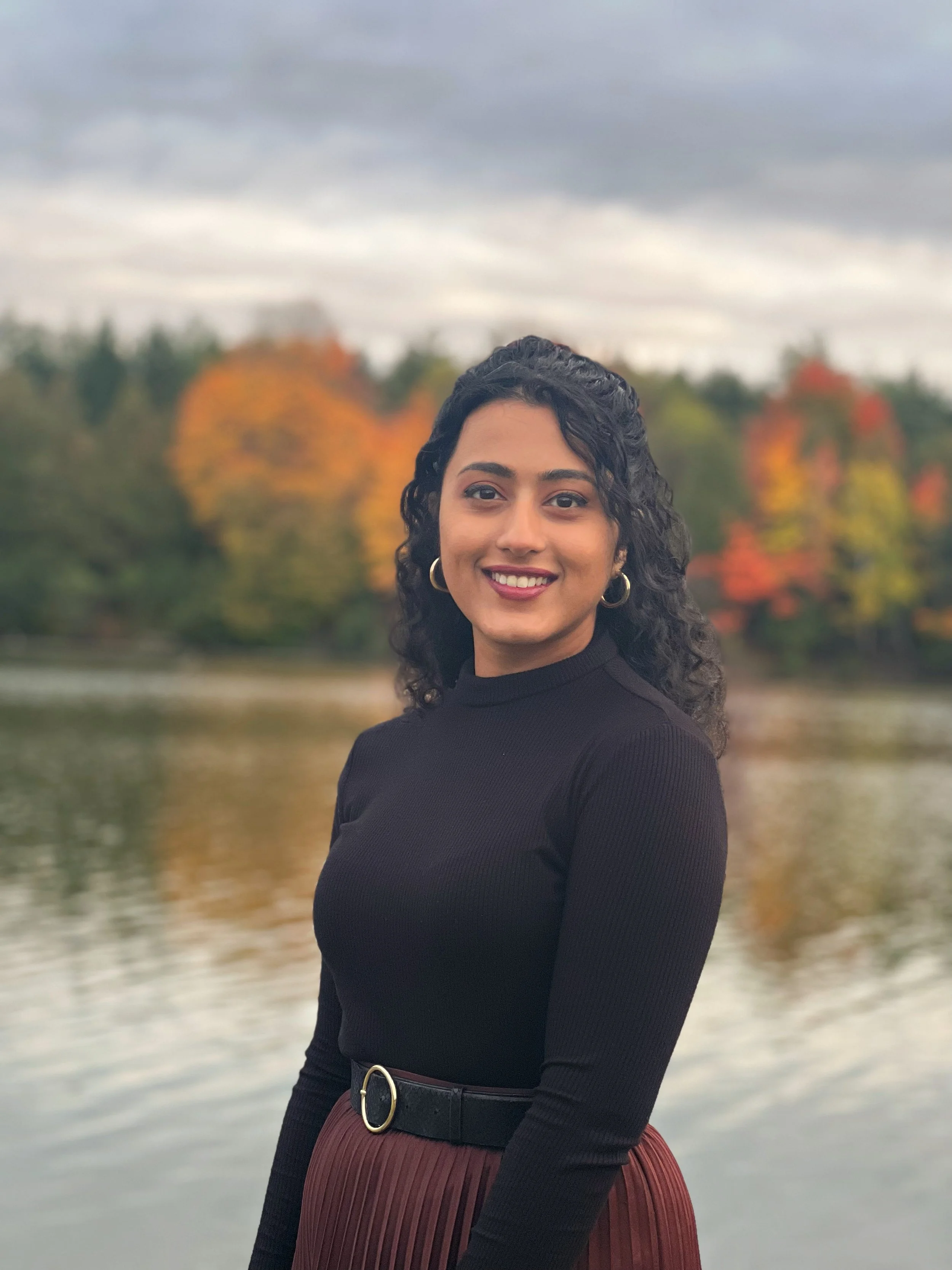 A woman with curly dark hair smiling outdoors near a body of water with trees displaying fall colors in the background.