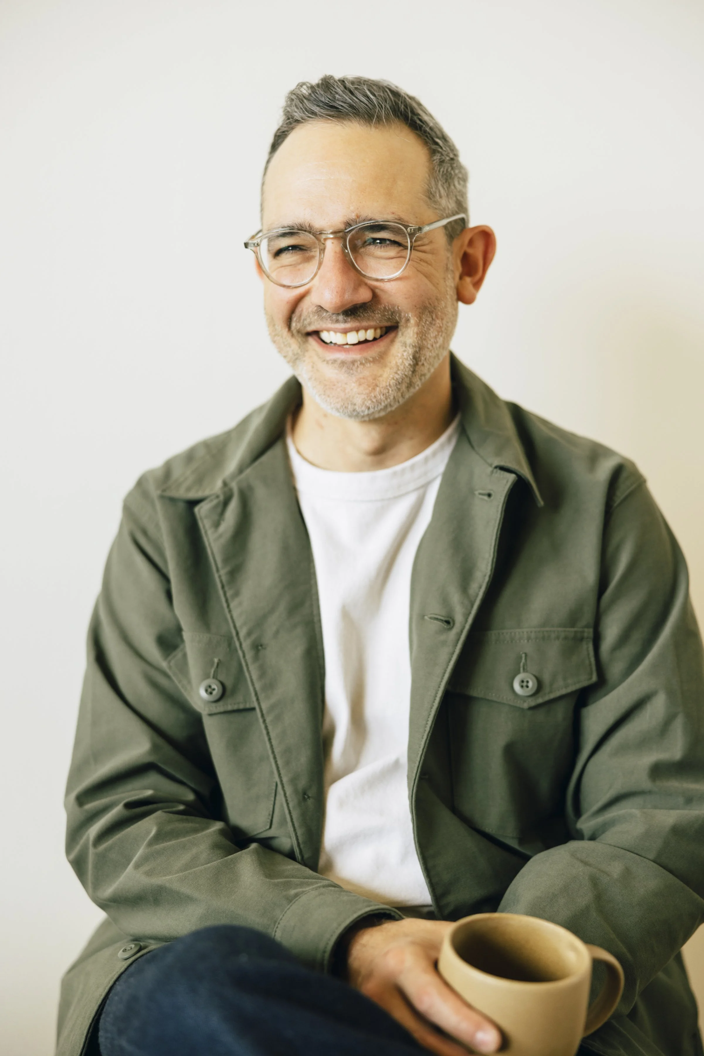 A smiling middle-aged man with glasses, gray hair, and a beard, wearing a white T-shirt and an olive-green jacket, sitting casually while holding a beige coffee mug.