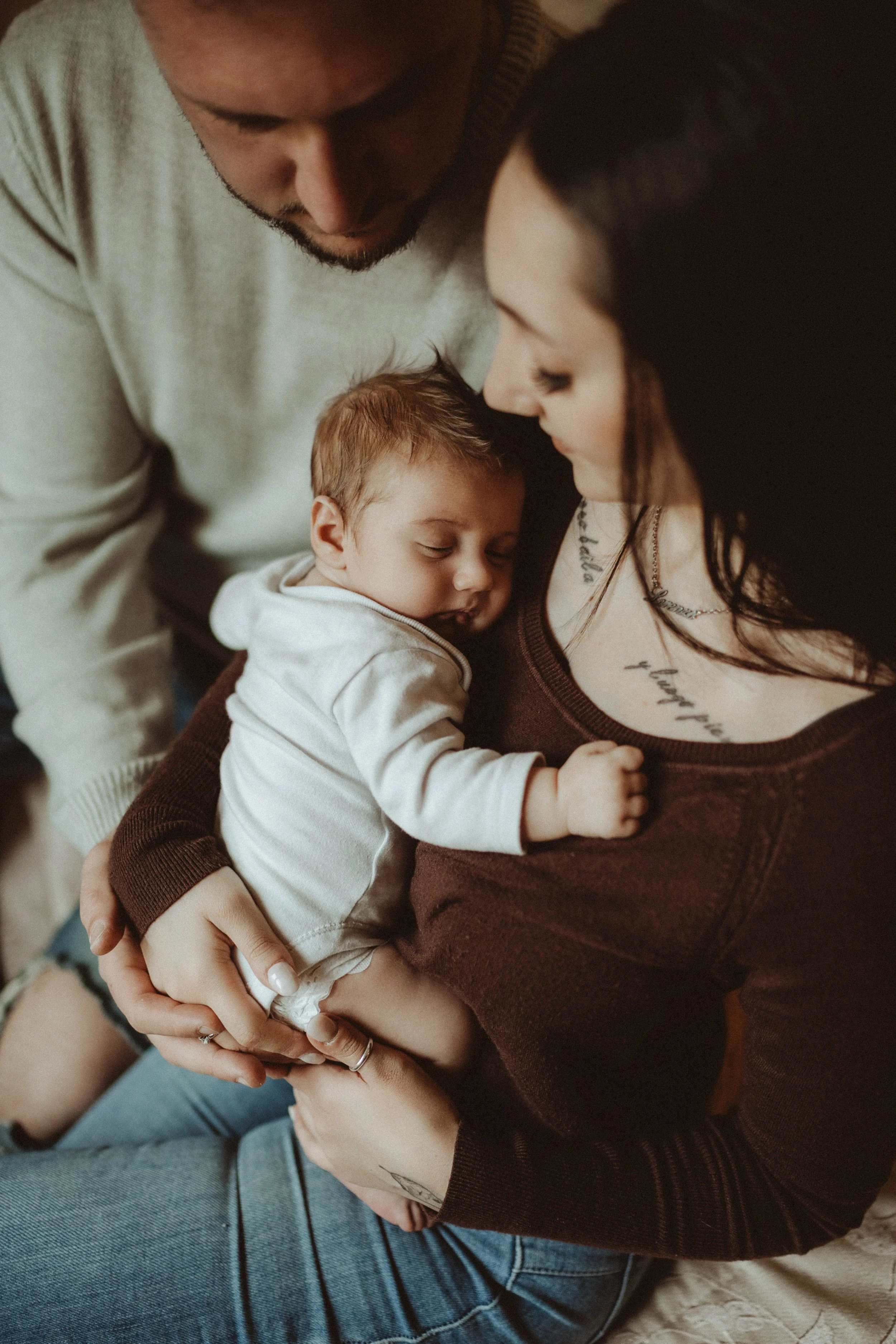 Una coppia con un bambino tra le braccia, abbracciati e sorridenti, in un momento di tenerezza domestica.