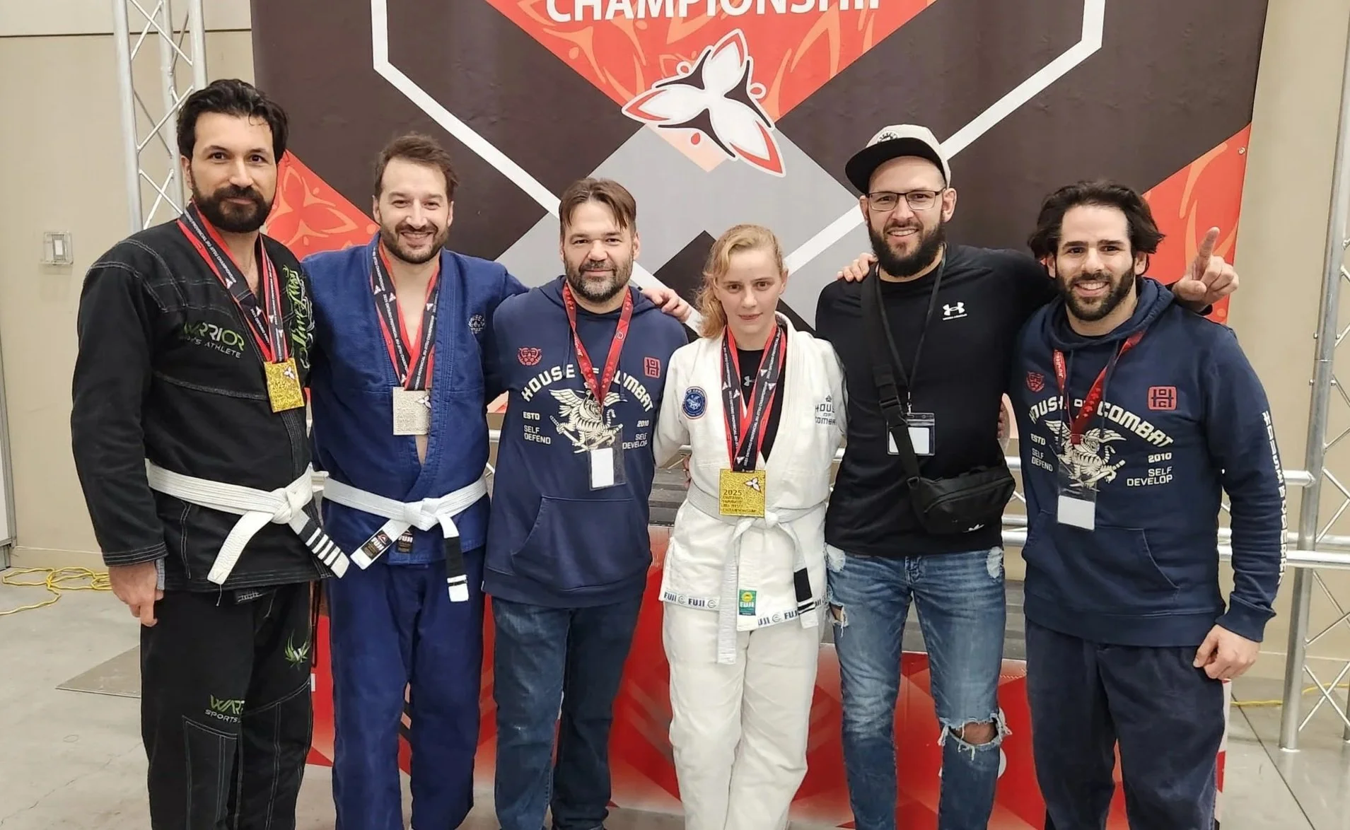 Six people, five men and one woman, standing together in front of a martial arts tournament banner, wearing martial arts gis and medals.
