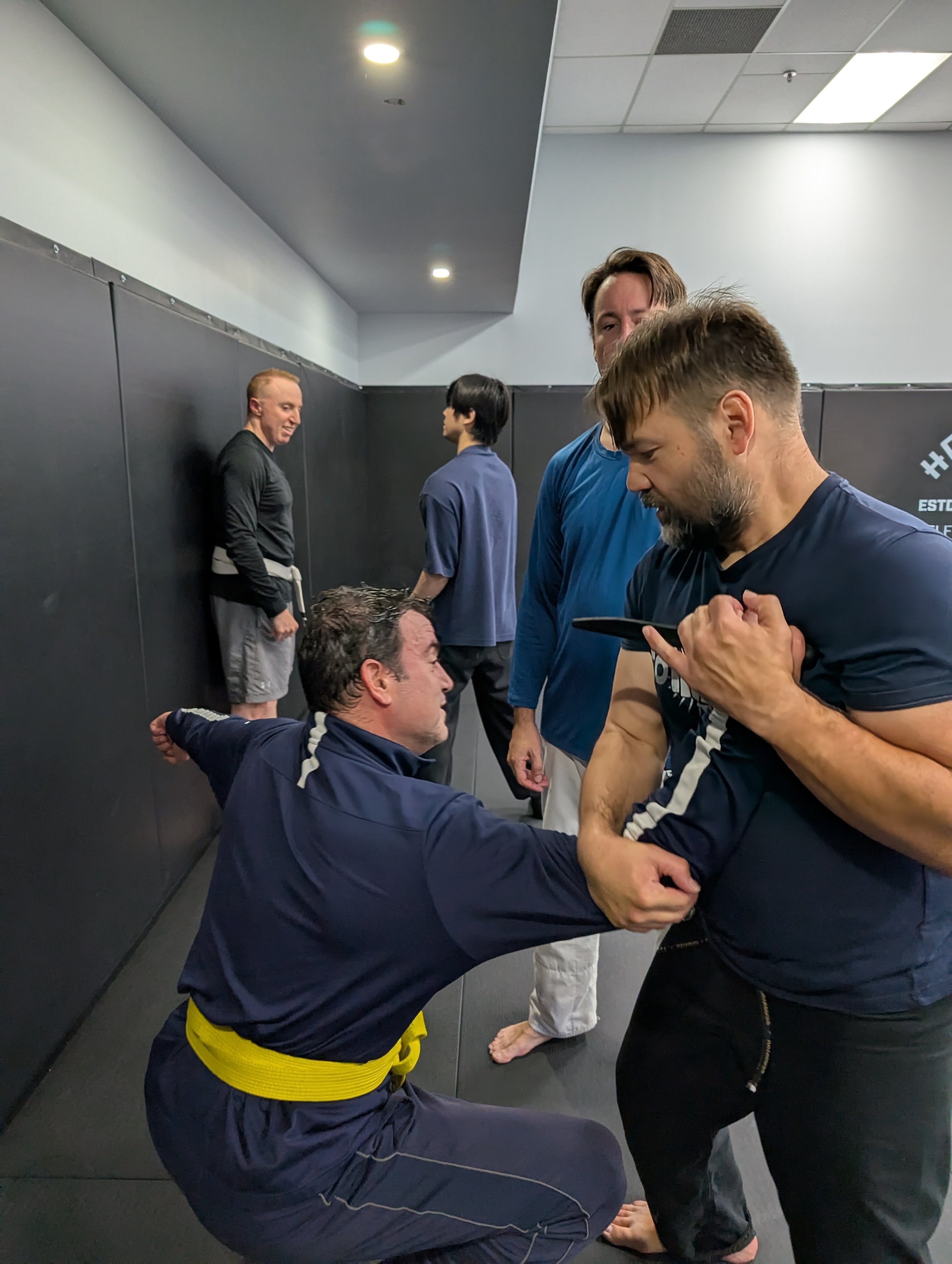 Group of men practicing Krav Maga knife defence techniques in a training room with black mats and wall padding, one man wearing a yellow belt and three others in casual athletic clothing.