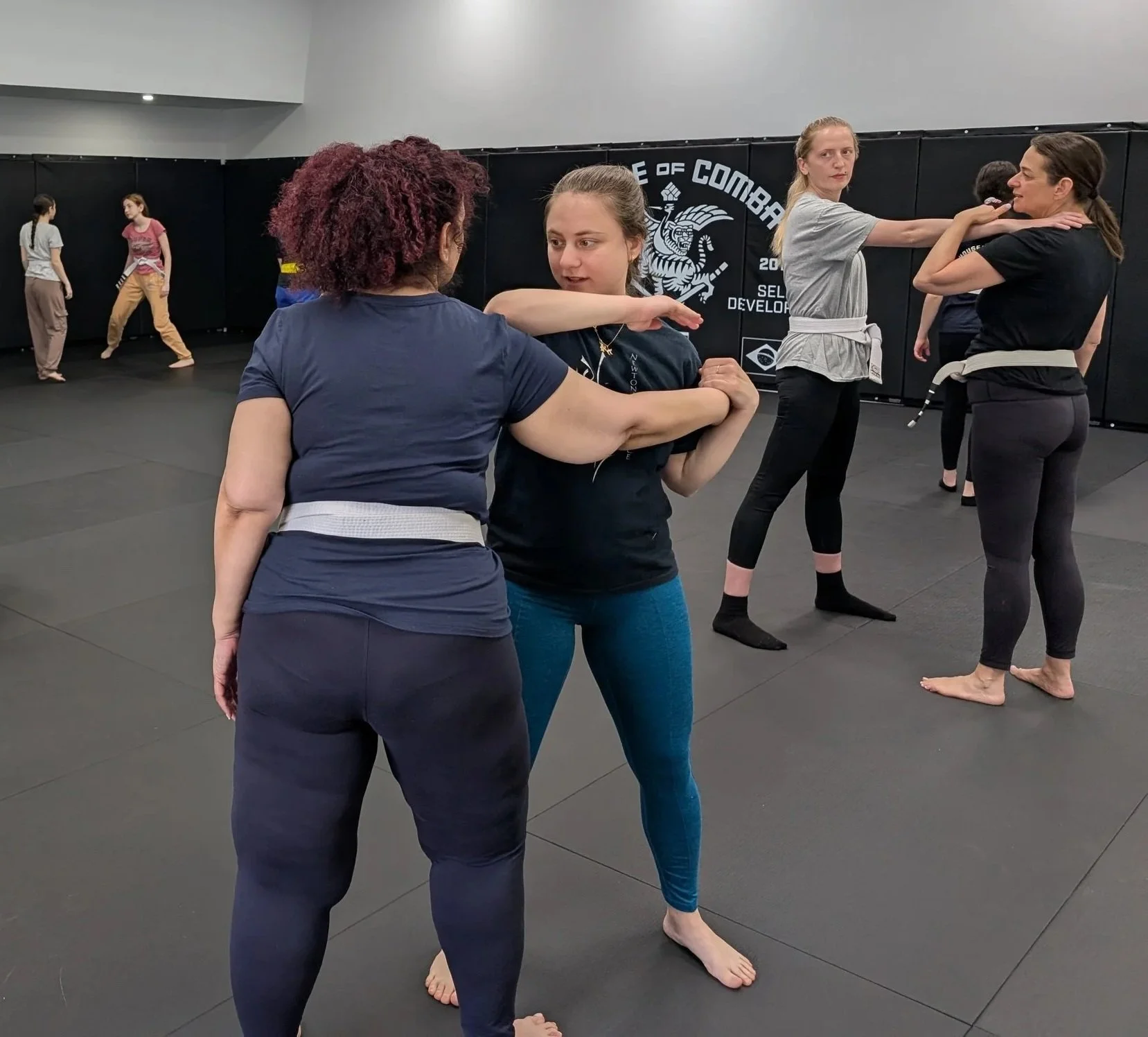 Two women practicing self defence techniques in a martial arts gym. One woman with curly red hair is demonstrating a move to another woman with dark hair tied back, while other women in the background spar or practice techniques.