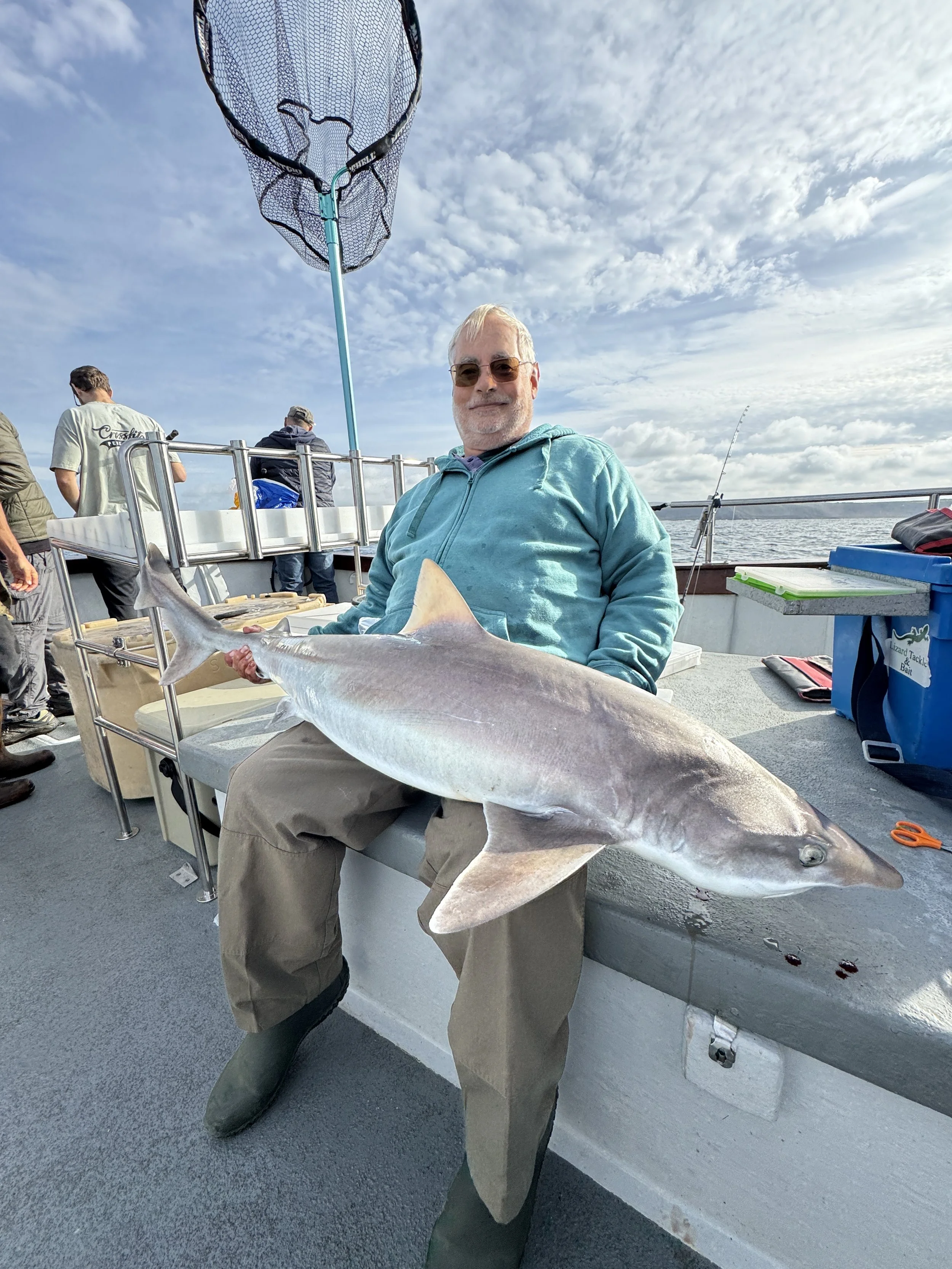 An elderly man sitting on a boat holding a large fish, with other fishermen and fishing equipment in the background, on a cloudy day.