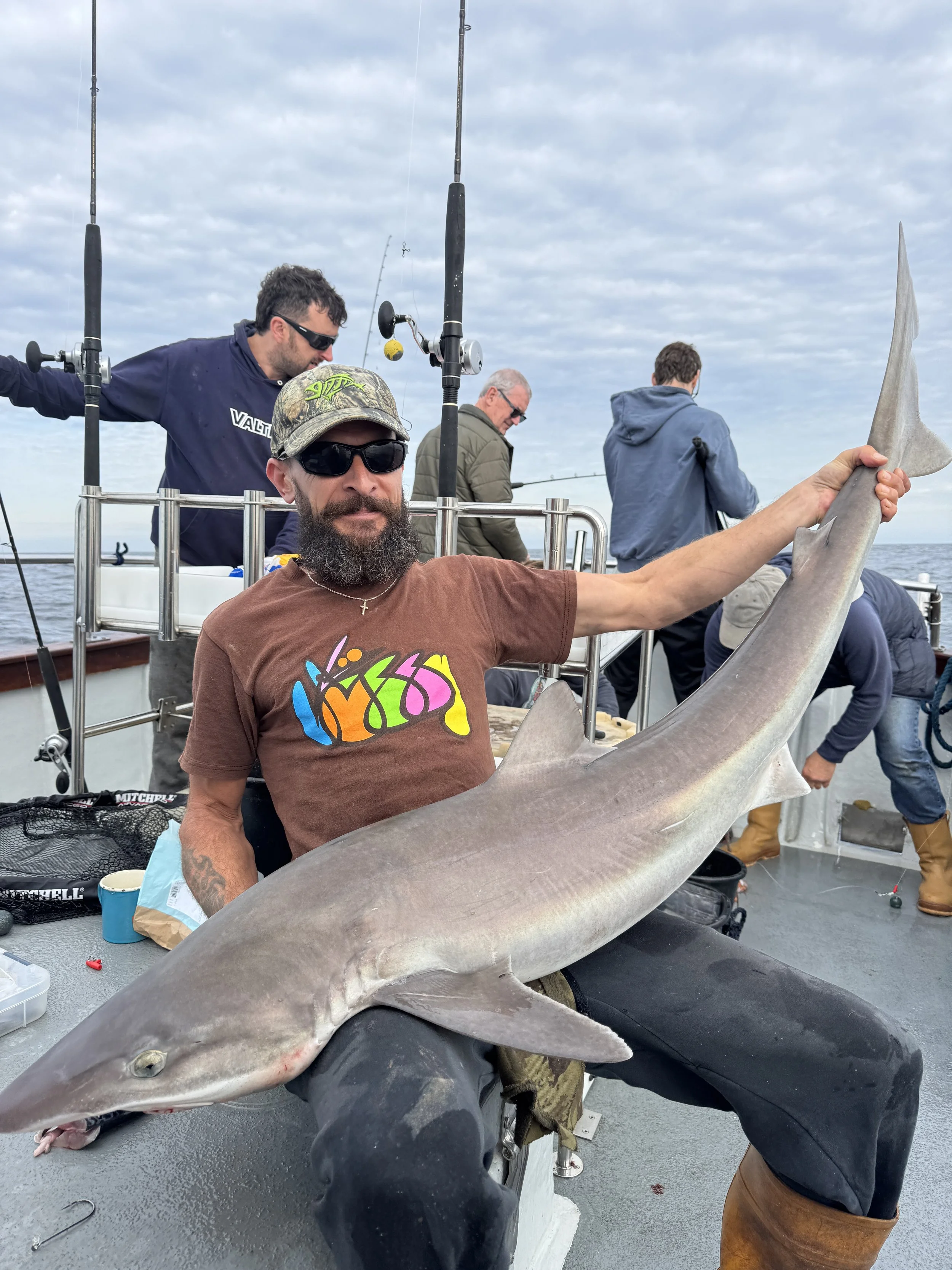 Man holding a large shark on a boat with other people fishing in the background.