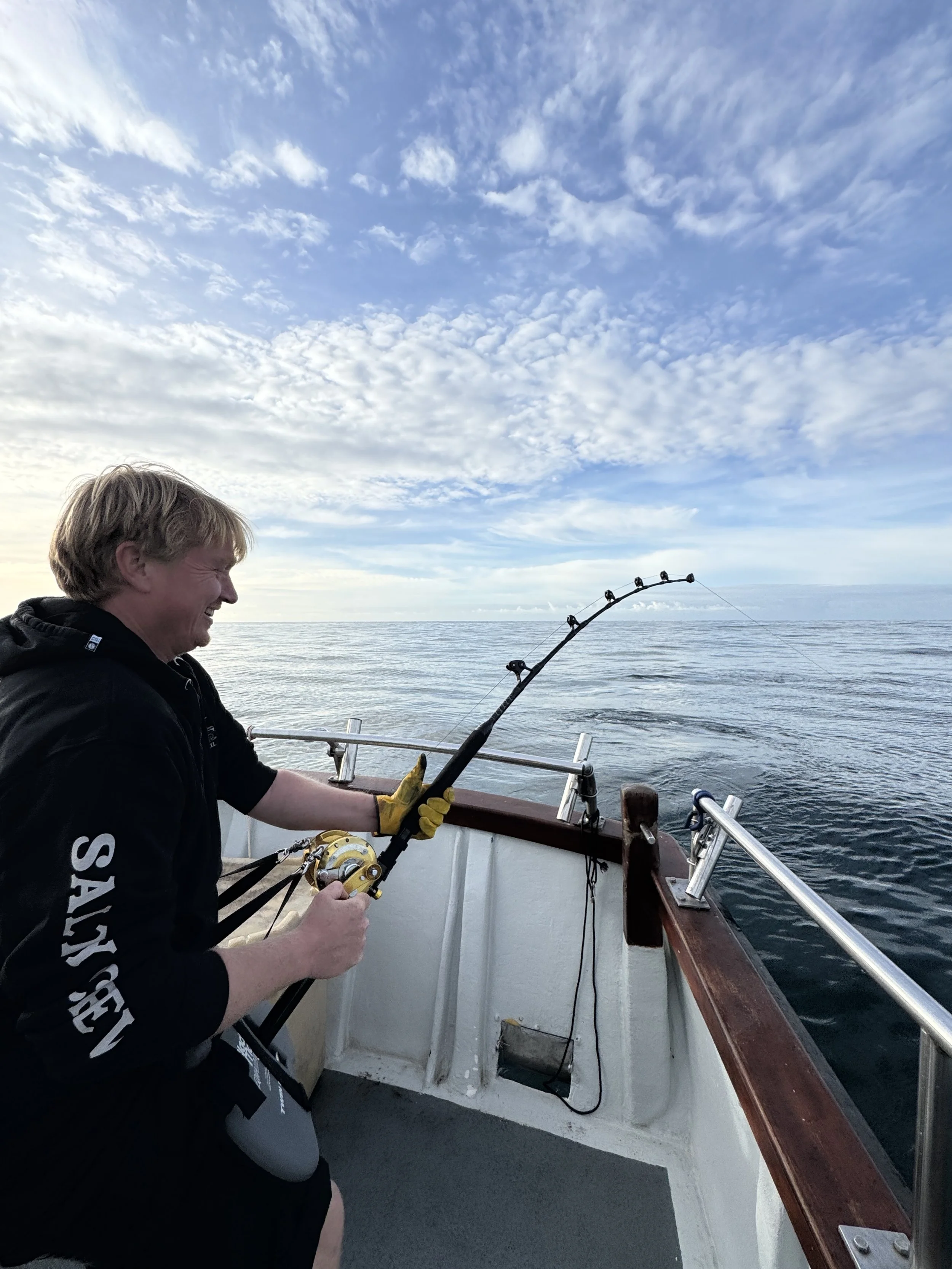 A man on a boat holding a fishing rod, smiling as he catches a fish near the water with a cloudy sky overhead.