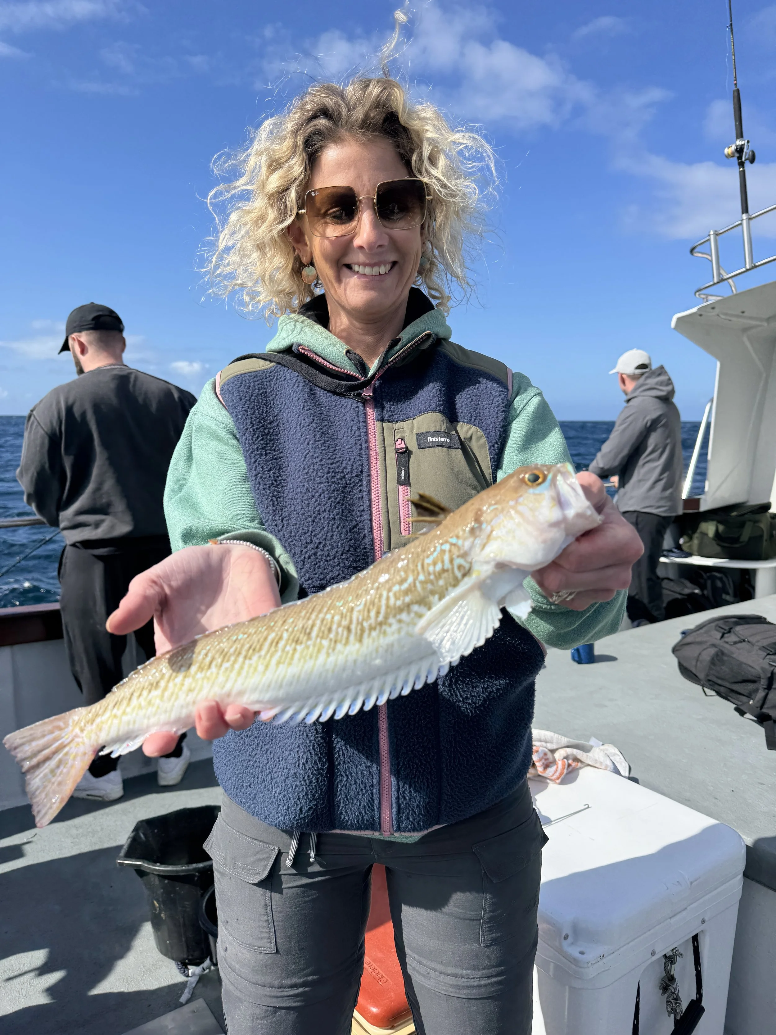 Woman with curly blonde hair wearing sunglasses and a colorful jacket on a boat holding a fish she caught. Two men are in the background, fishing or working on the boat, with the ocean and sky visible.