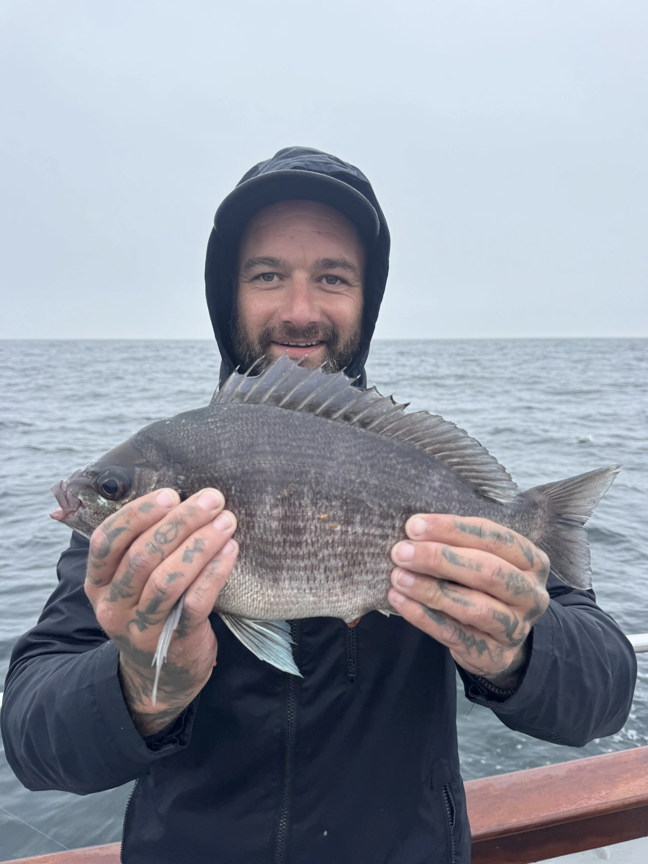 A man with a beard and tattoos holds a large fish on a boat, with the ocean in the background, under a cloudy sky.