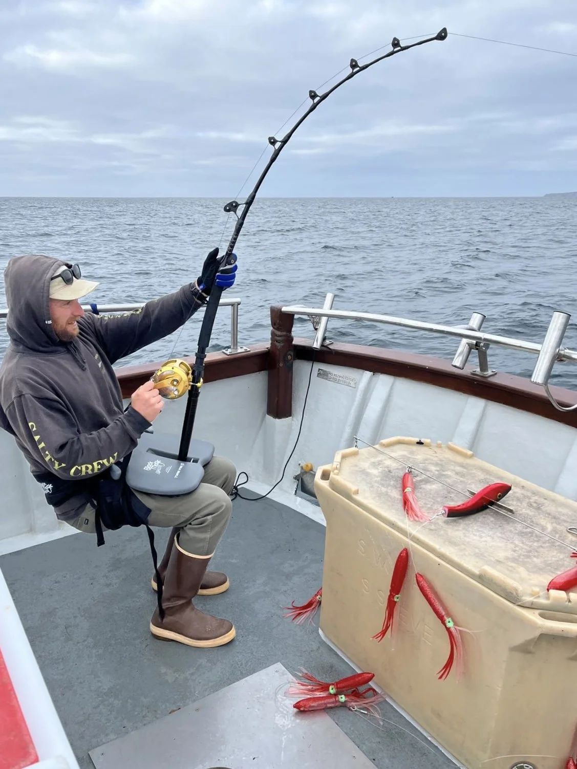A man on a boat fishing with a rod and reel, wearing a hoodie, gloves, and boots, with seabed markers attached to the fishing line. The ocean is visible in the background under a cloudy sky.