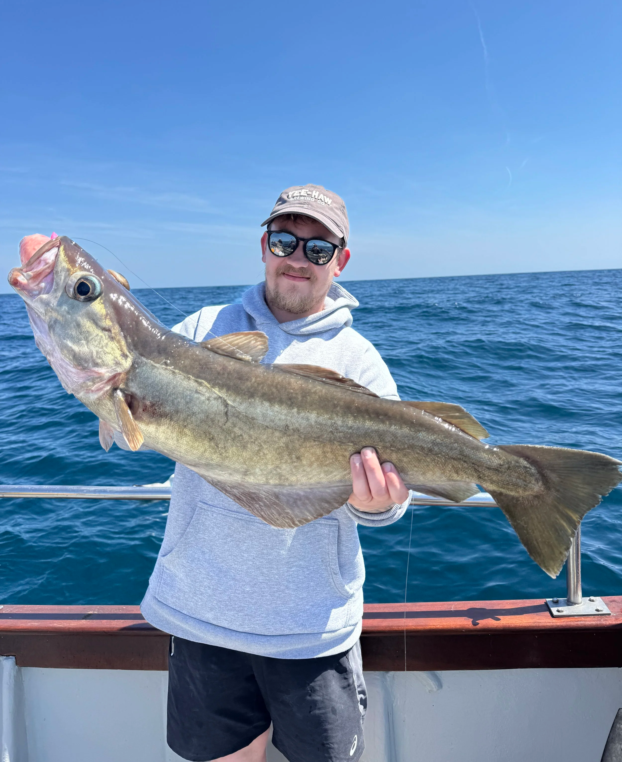 A man with sunglasses and a gray hoodie holding a large fish on a boat at sea under a clear blue sky.
