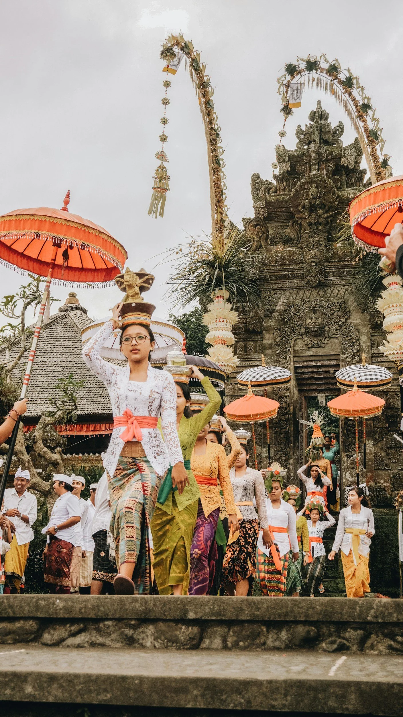 People dressed in traditional Balinese attire participating in a cultural procession at a historic temple, carrying umbrellas and offerings.