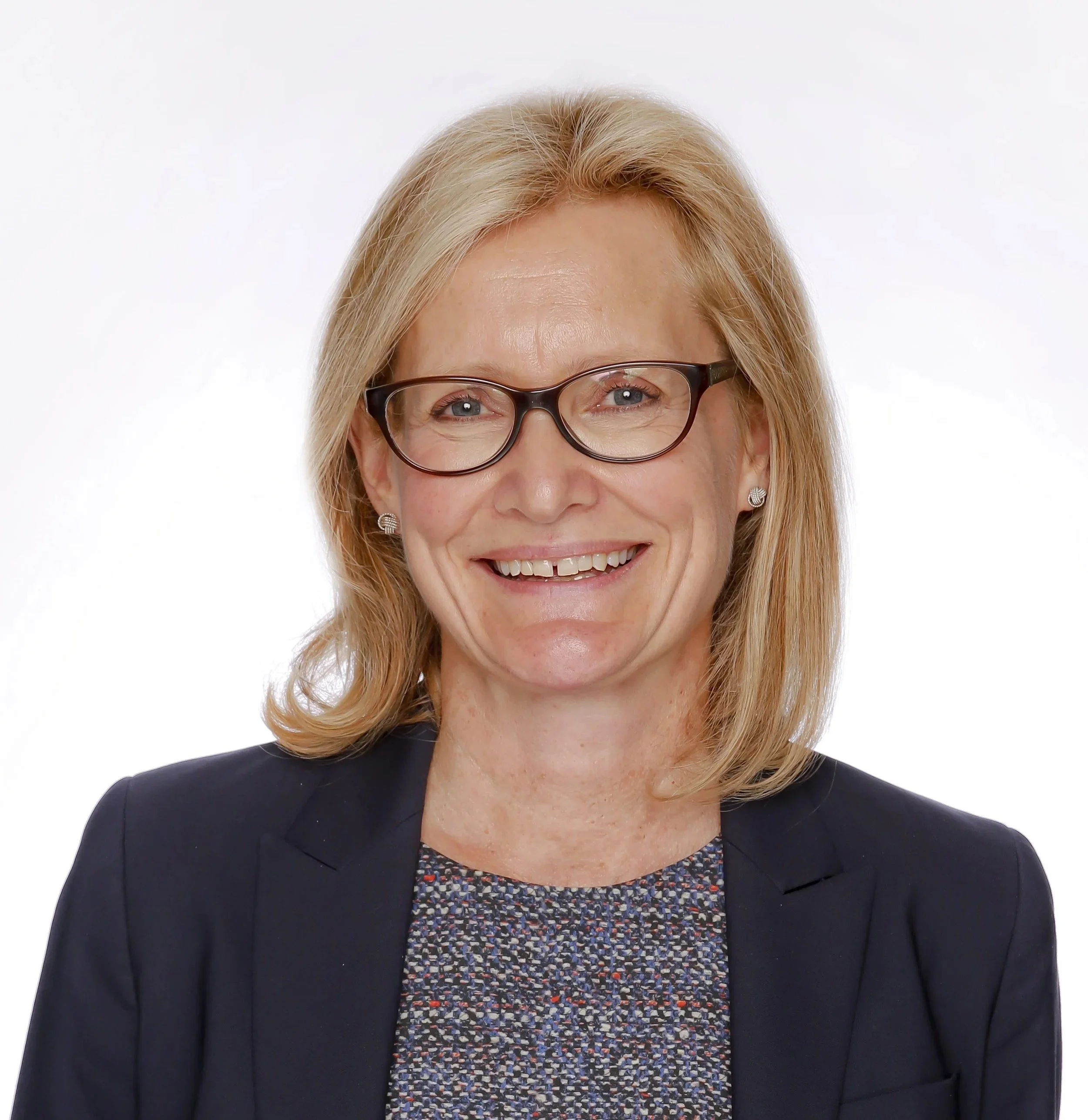 Close-up of a smiling woman with light skin, blonde hair, wearing black-rimmed glasses, pearl earrings, a dark blazer, and a colorful patterned blouse against a white background.