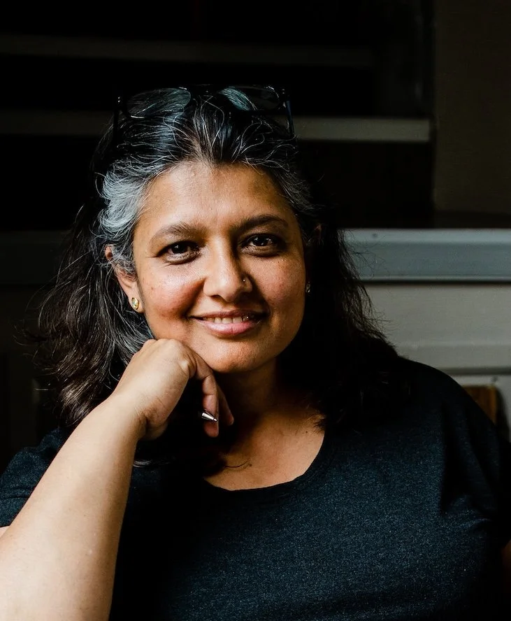A woman with dark hair and glasses on her head, smiling and resting her chin on her hand, wearing a black shirt.