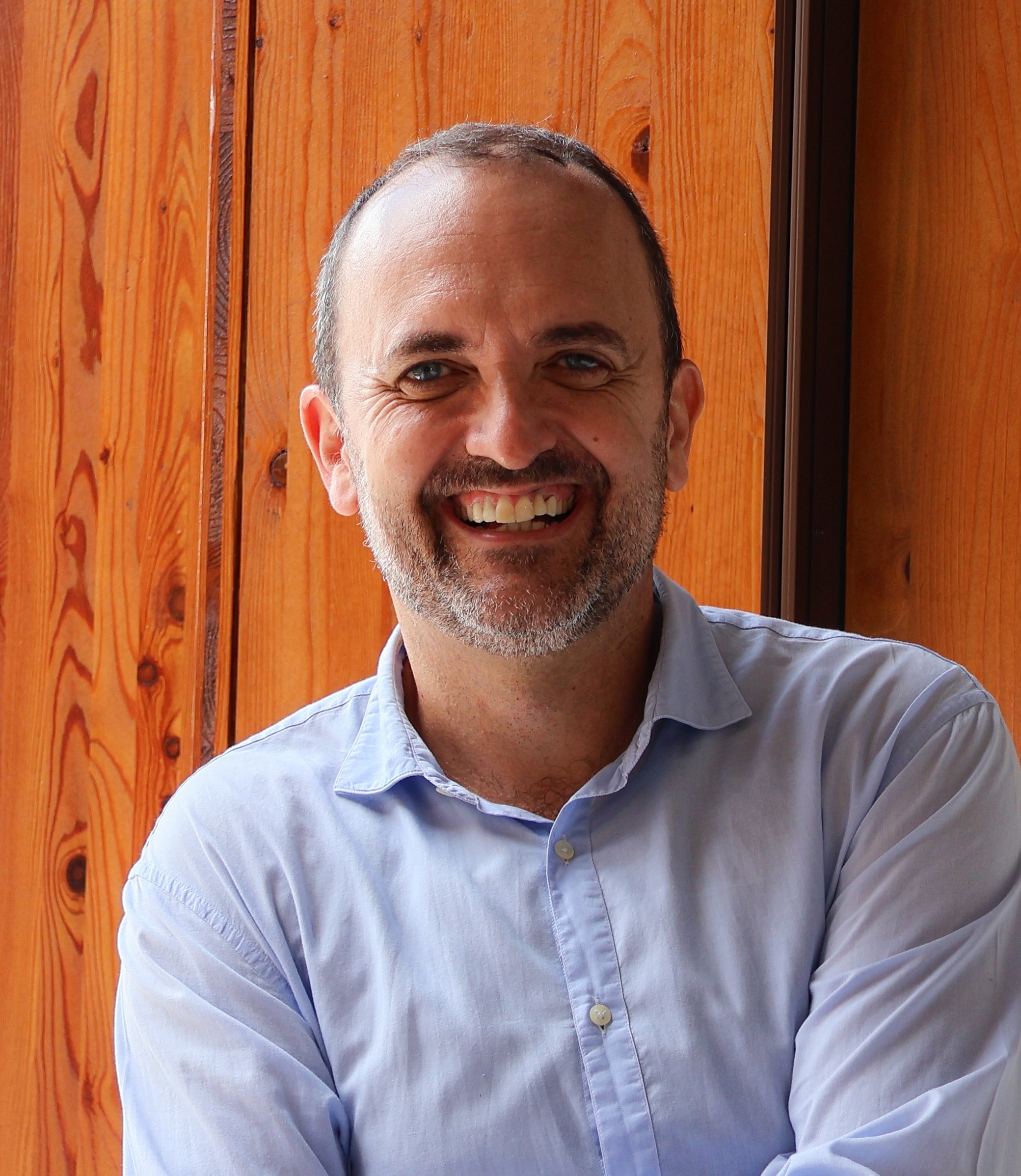 A smiling man with light skin, short dark hair, and a beard wearing a light blue button-up shirt, sitting in front of a wooden wall.