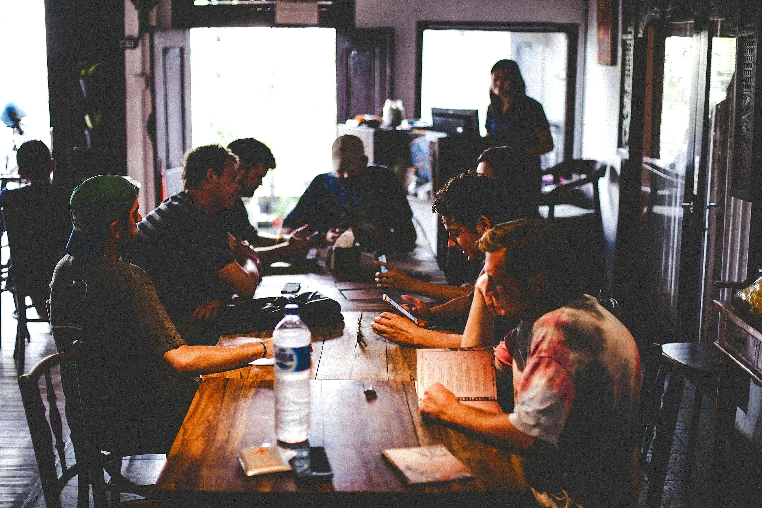 A group of people sitting around a large wooden table in a dimly lit room, with some looking at their phones and a woman standing at the back near a computer, possibly giving a presentation or leading a discussion.