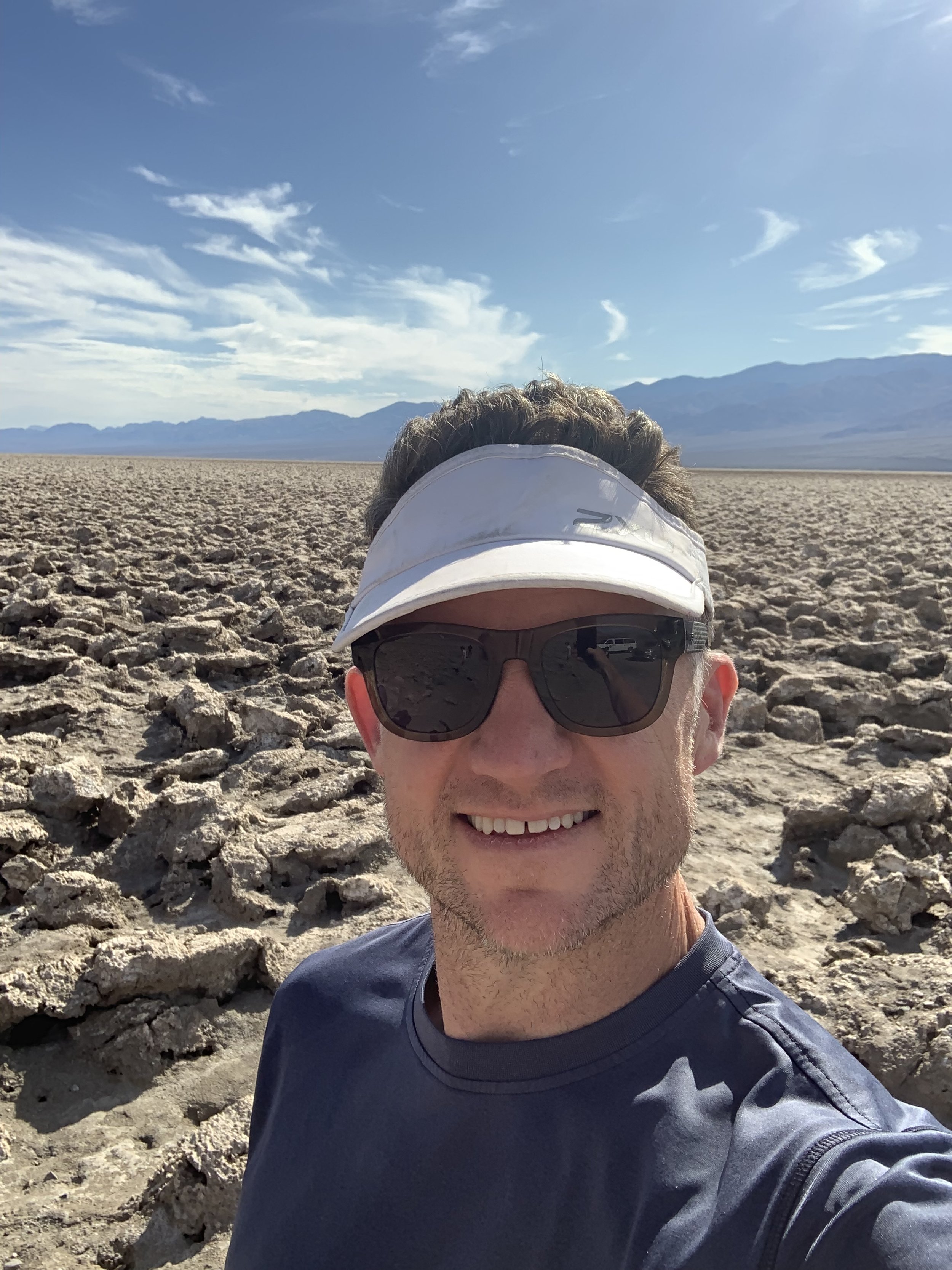 Rob wearing sunglasses and a visor taking a selfie in a dry, rocky desert landscape with distant mountains and a partly cloudy sky.