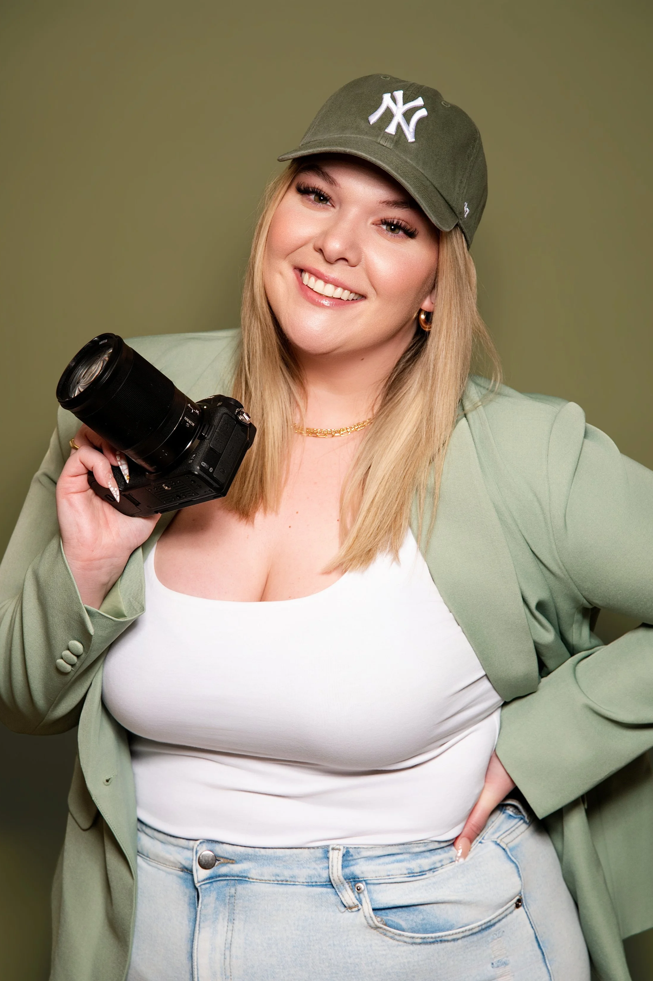A smiling woman wearing a green New York Yankees baseball cap, light green blazer, white tank top, and light-wash jeans, holding a camera against a plain olive-green background.