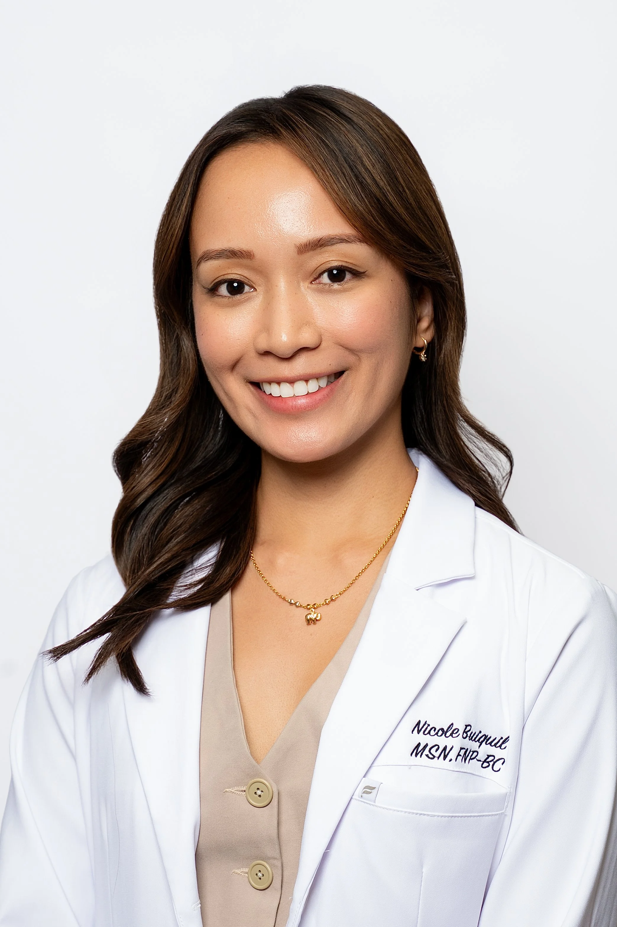 Smiling woman with long brown hair, wearing a white coat with her name and credentials embroidered, a beige top with buttons, and gold jewelry, standing against a plain white background.