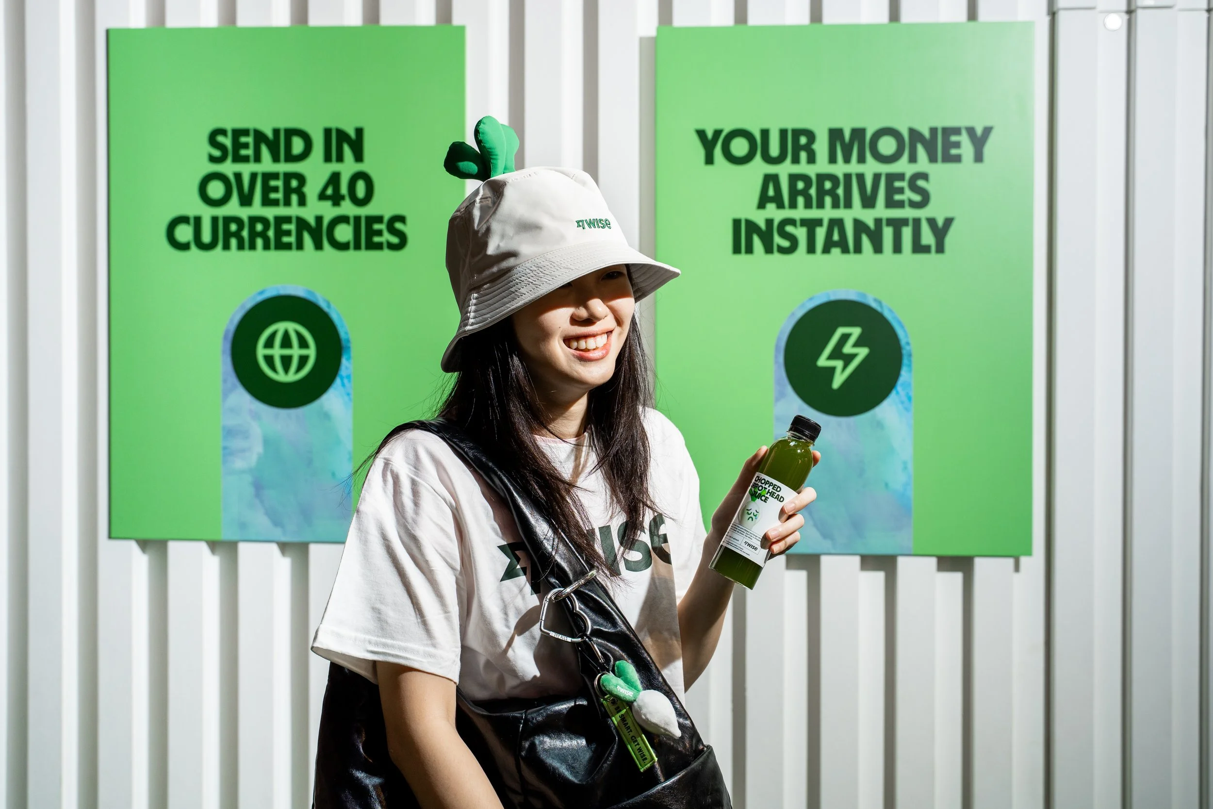 A woman wearing a bucket hat and a white t-shirt holding a green juice bottle, standing in front of two green advertisements for a currency exchange company called Wise, with a globe icon and a lightning bolt icon.