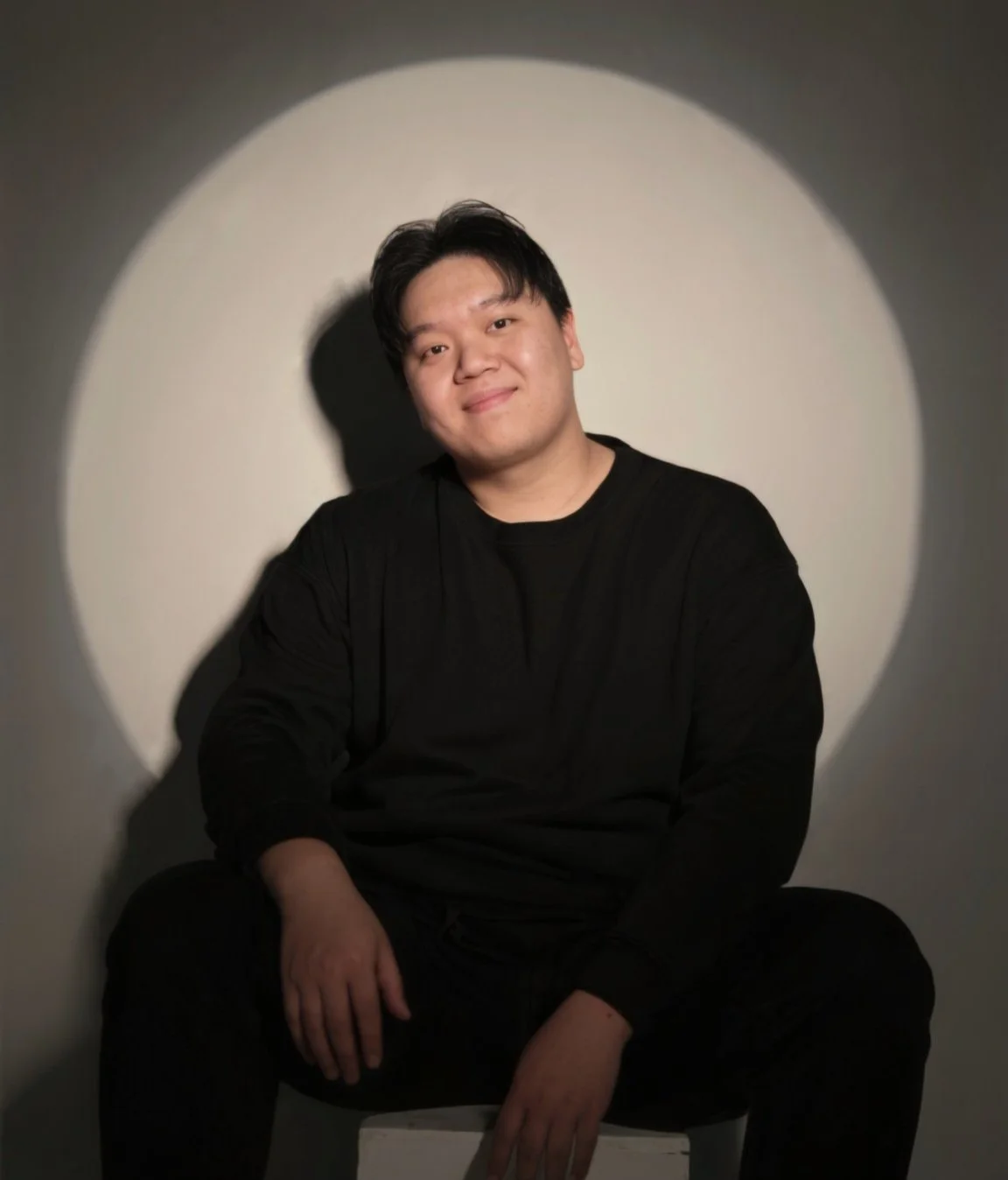 A young man sitting on a chair, wearing a black shirt and smiling, illuminated by a spotlight creating a circular light on the wall behind him.