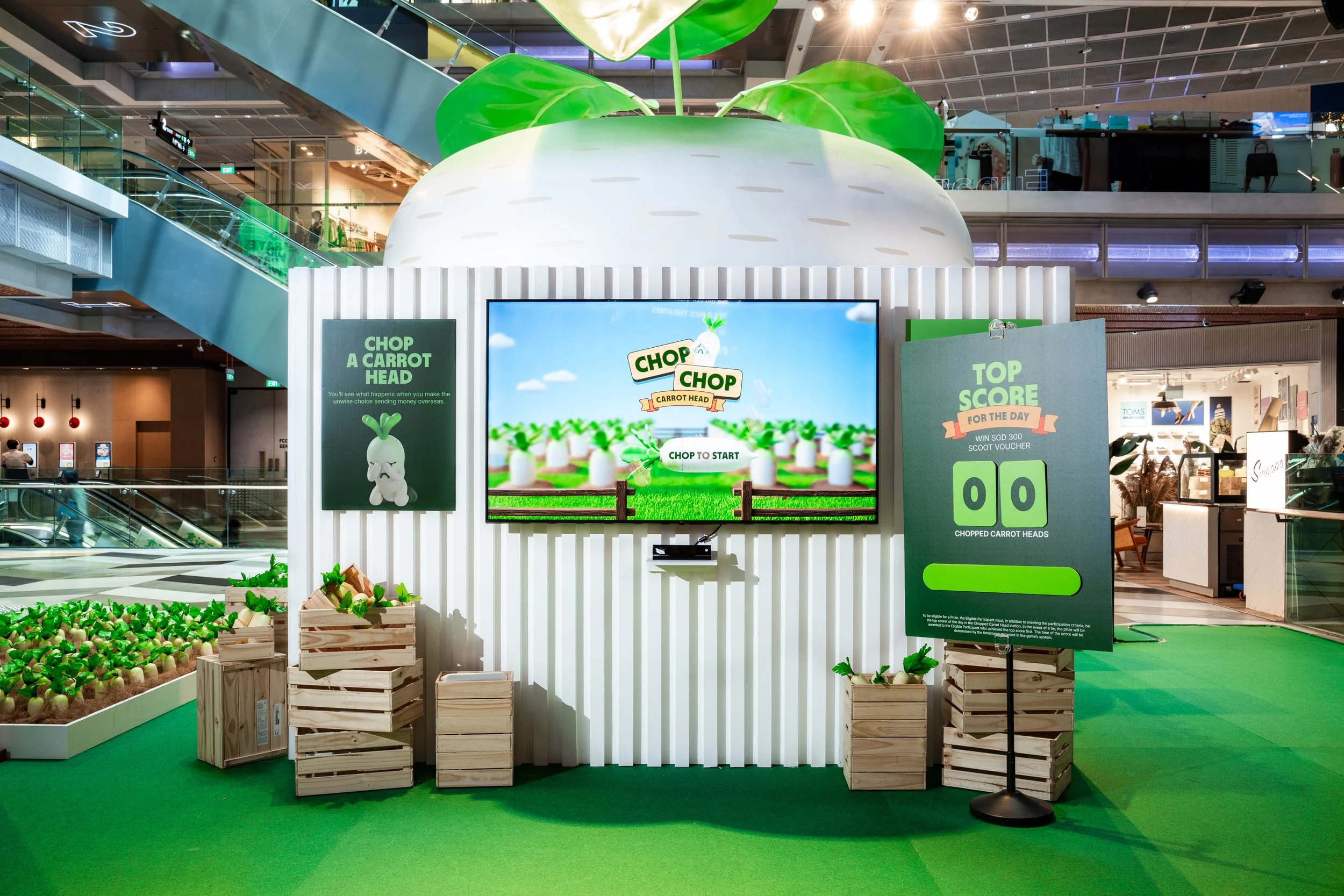 Indoor shopping mall kiosk with a large, decorative carrot head, promoting a game called 'Chop a Carrot Head.' The kiosk has bright green and white colors, with digital screens and signs about the game. There are small crates filled with green and white toy carrots and a small animated carrot character on the sign.