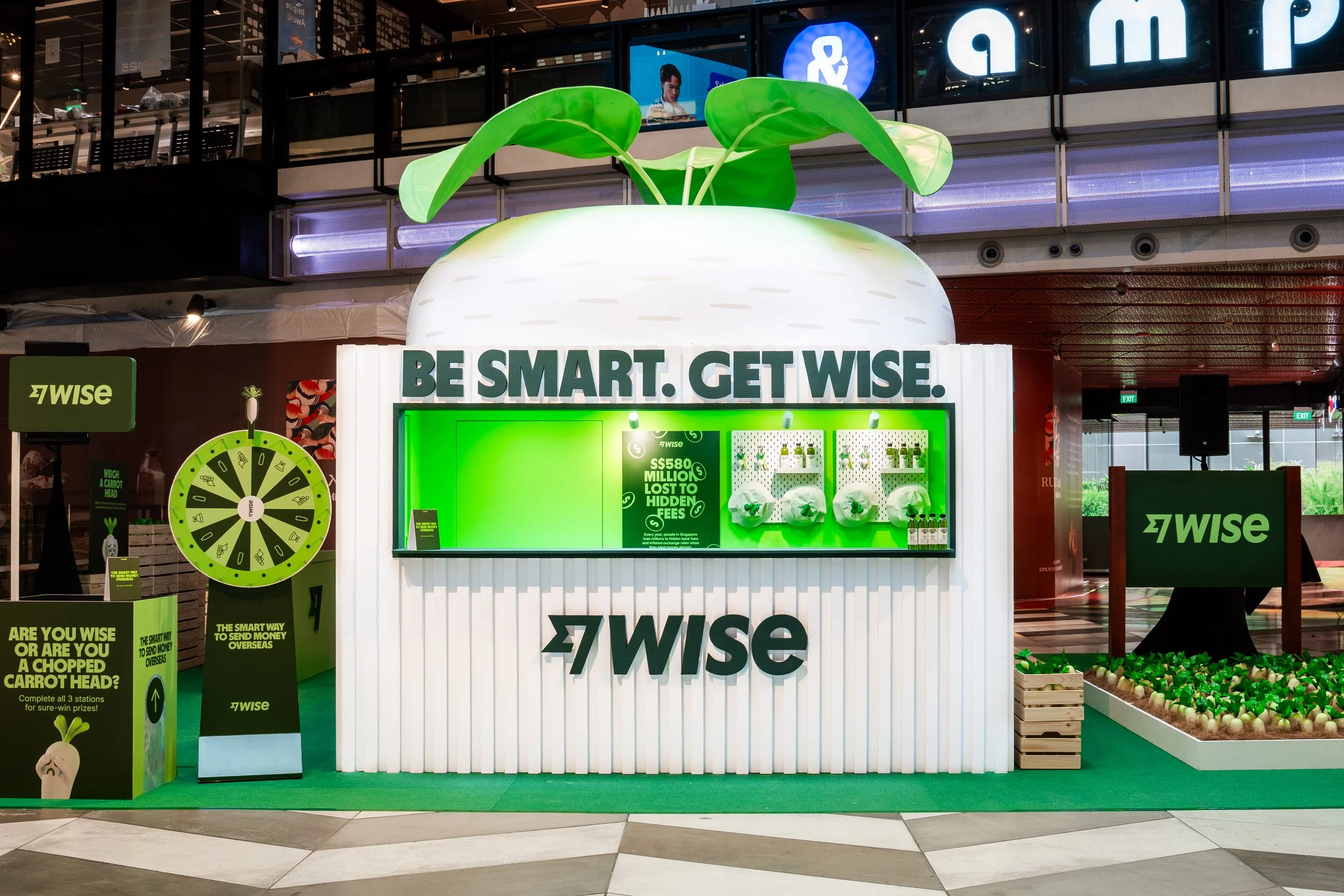 Bright green and white promotional booth for Wise, featuring vegetables like radishes and a large radish-shaped structure with leaves on top. The booth has the slogan 'Be Smart. Get Wise.' and informational posters about sending money overseas, with additional green-themed signage and a prize wheel.