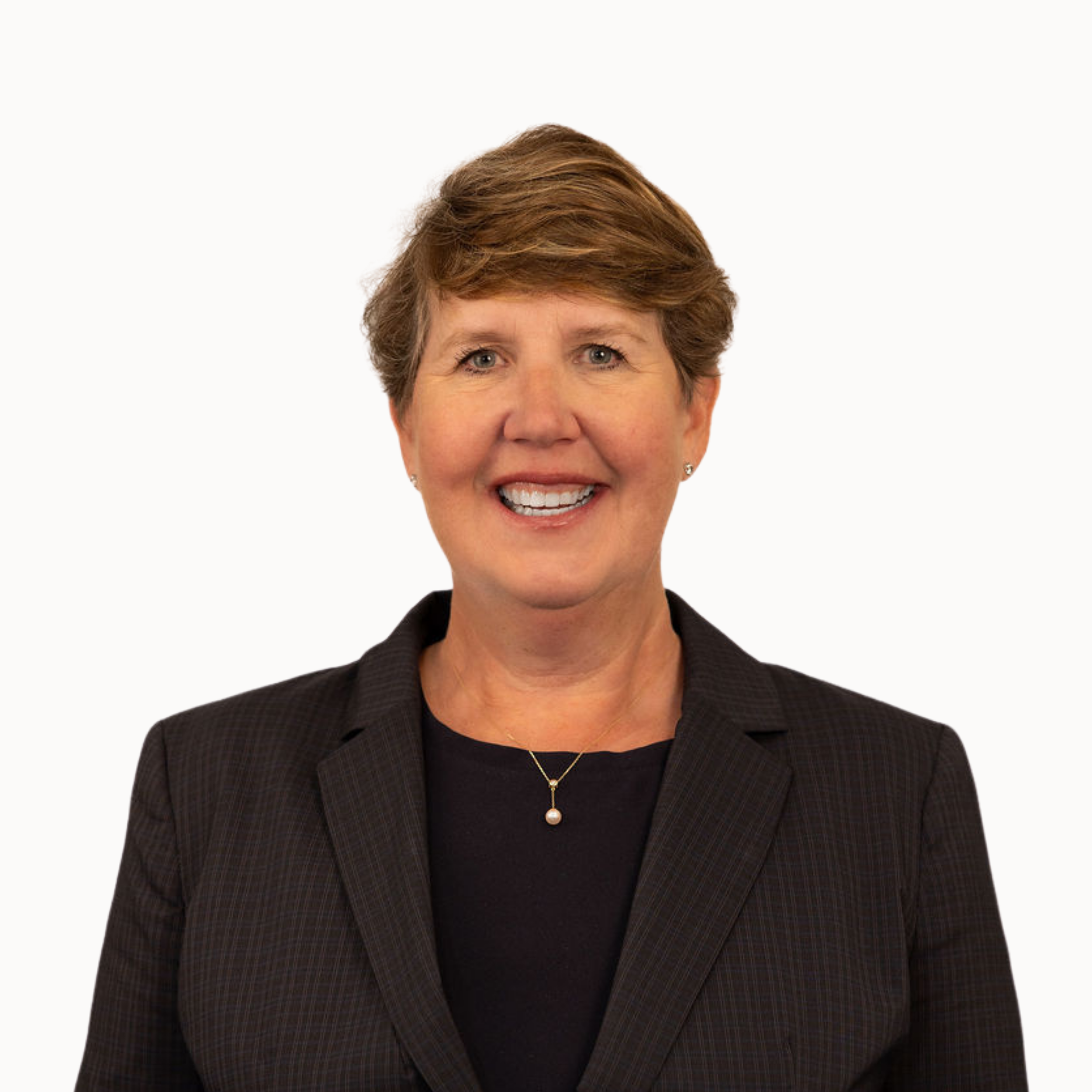 A portrait of a smiling woman with short reddish-brown hair, wearing pearl earrings, a black top, a gold necklace, and a white blazer with black stripes, against a dark gray background.