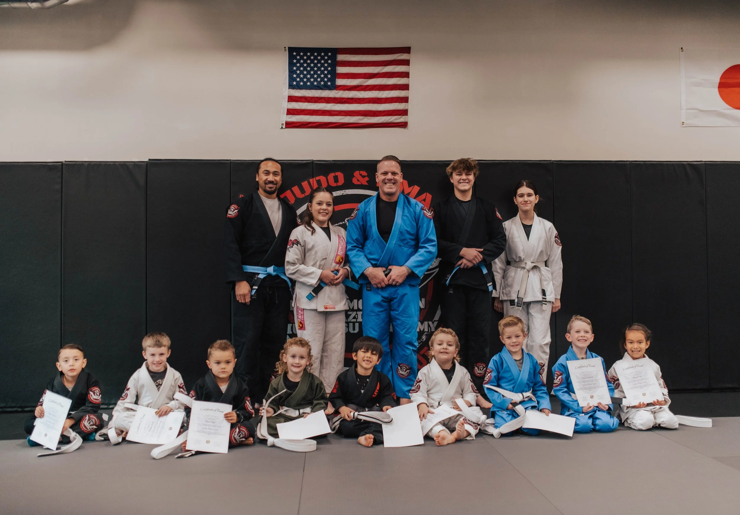 A martial arts instructor demonstrating a technique with a foam stick to a group of children in gi uniforms at a martial arts dojo.