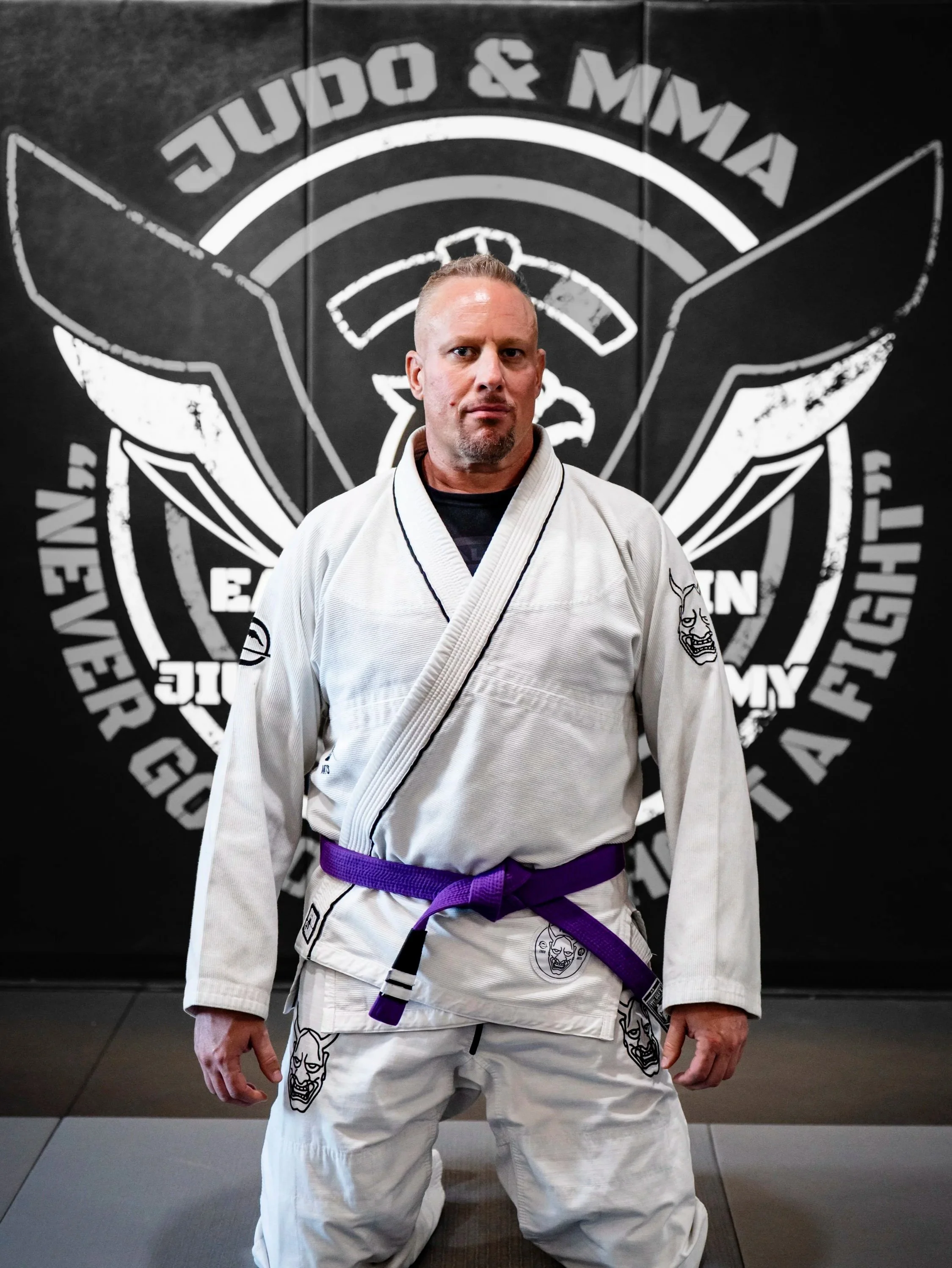 A man in a white Brazilian Jiu-Jitsu gi with a purple belt, standing in front of a Judo & MMA gym logo on the wall.