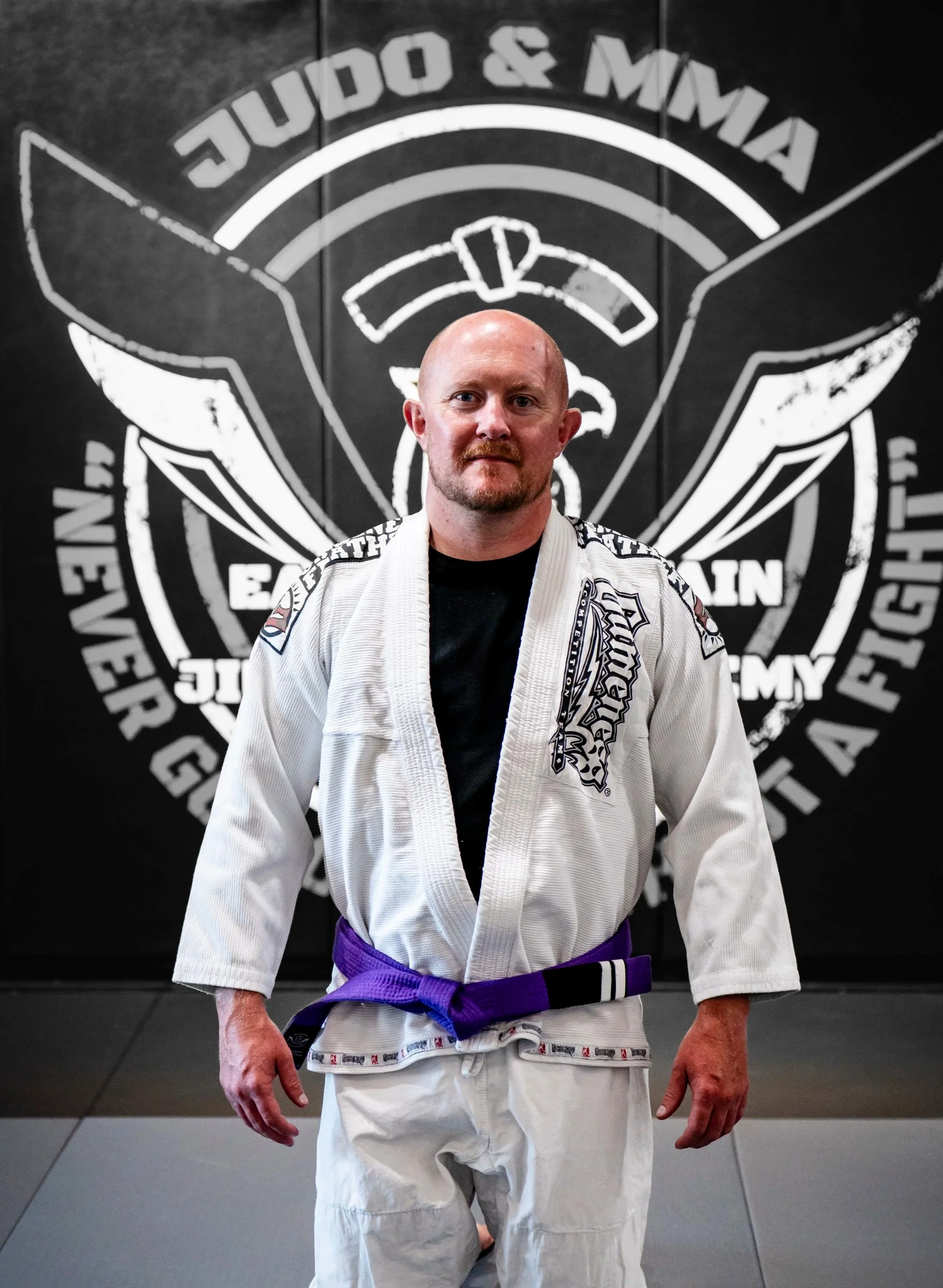 Bald man with a beard standing in a Brazilian Jiu-Jitsu gym, wearing a white gi with a purple belt, in front of a black wall with a large martial arts logo.
