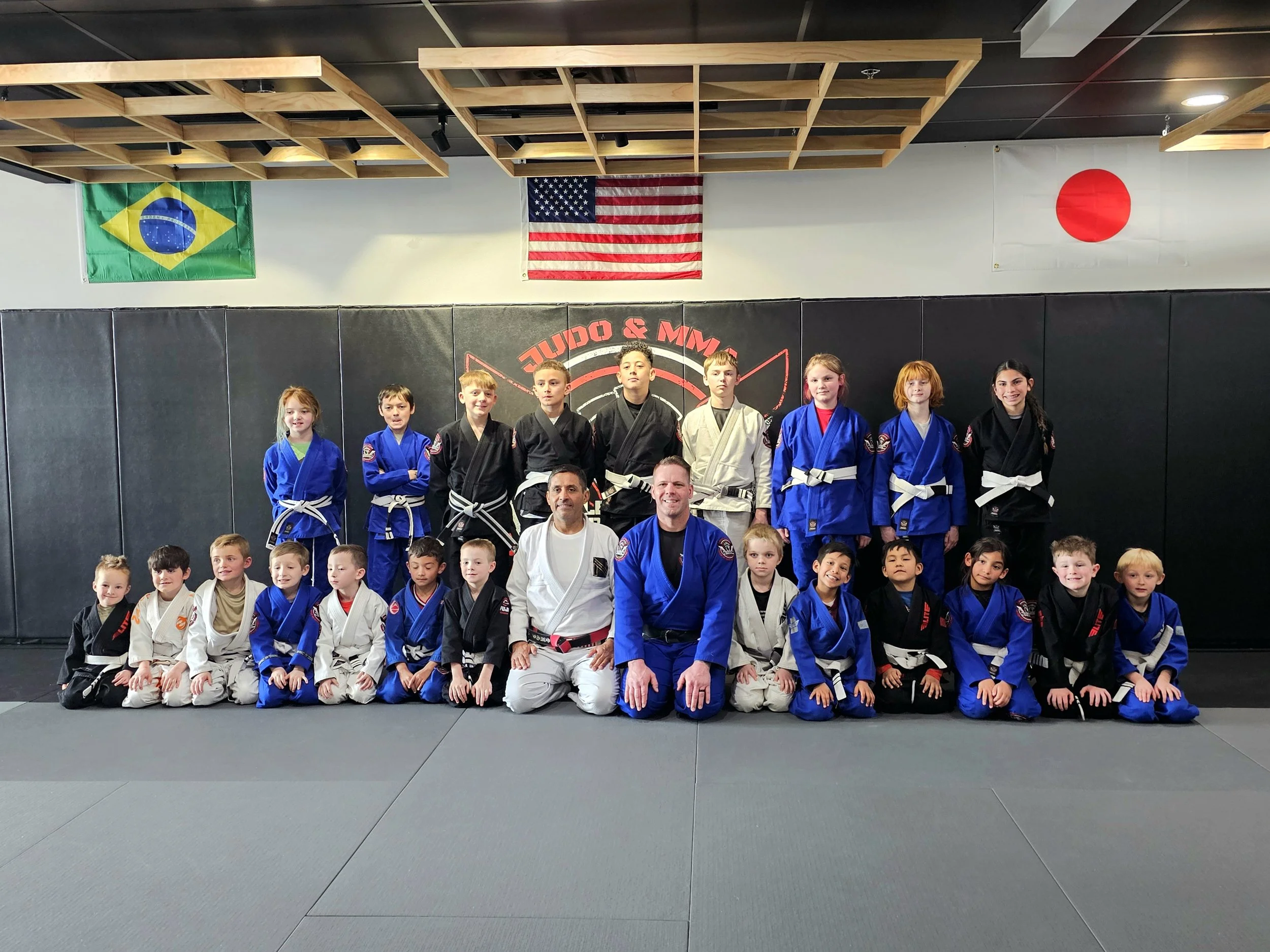 Four children practicing Brazilian Jiu-Jitsu in a gym, two grappling in the foreground and two standing in the background, with a digital clock showing -9:41 on the wall.