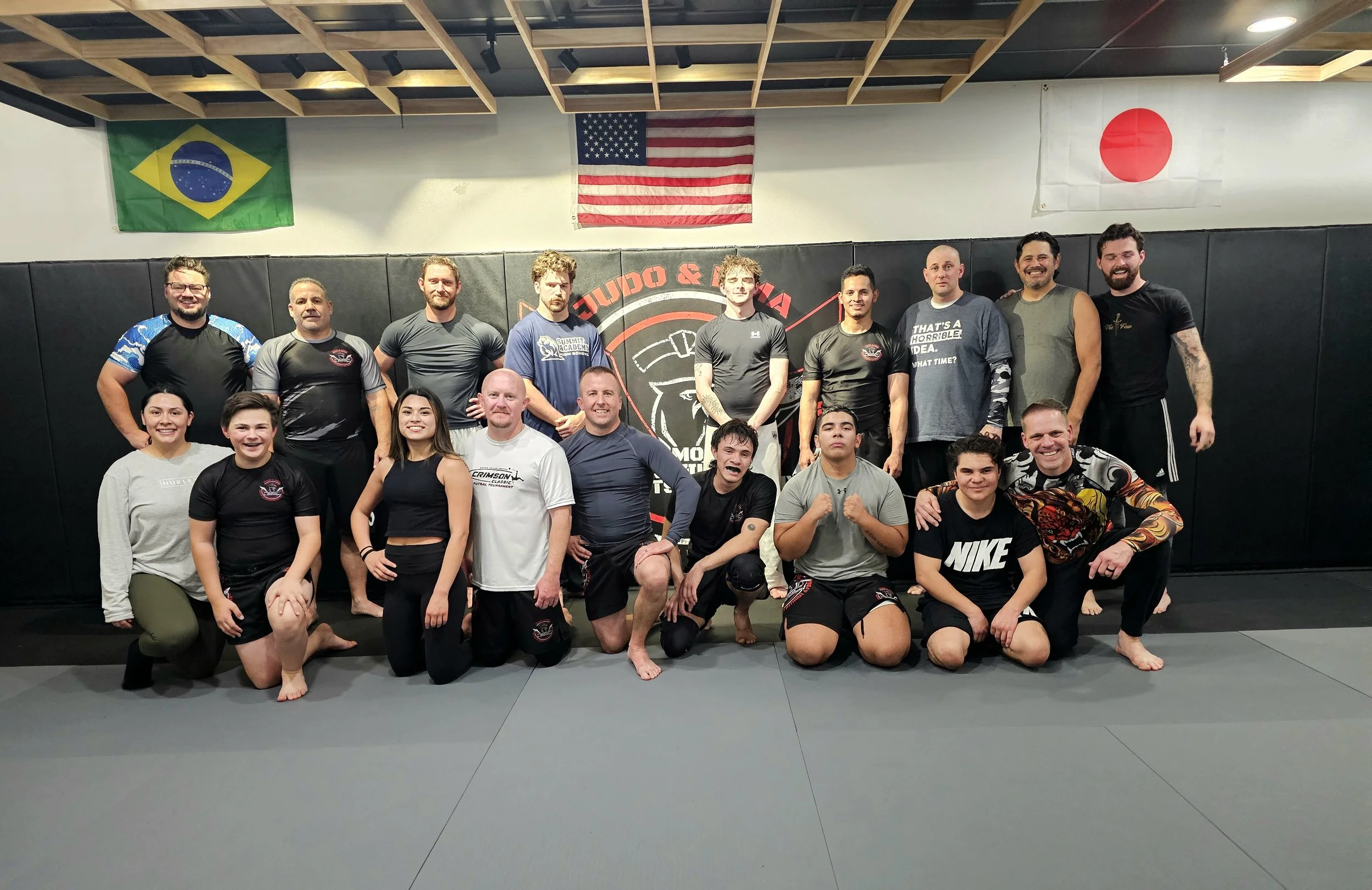 Group of people in mixed martial arts training gear, posing together in a martial arts gym with flags of Brazil, the United States, and Japan hanging above them.