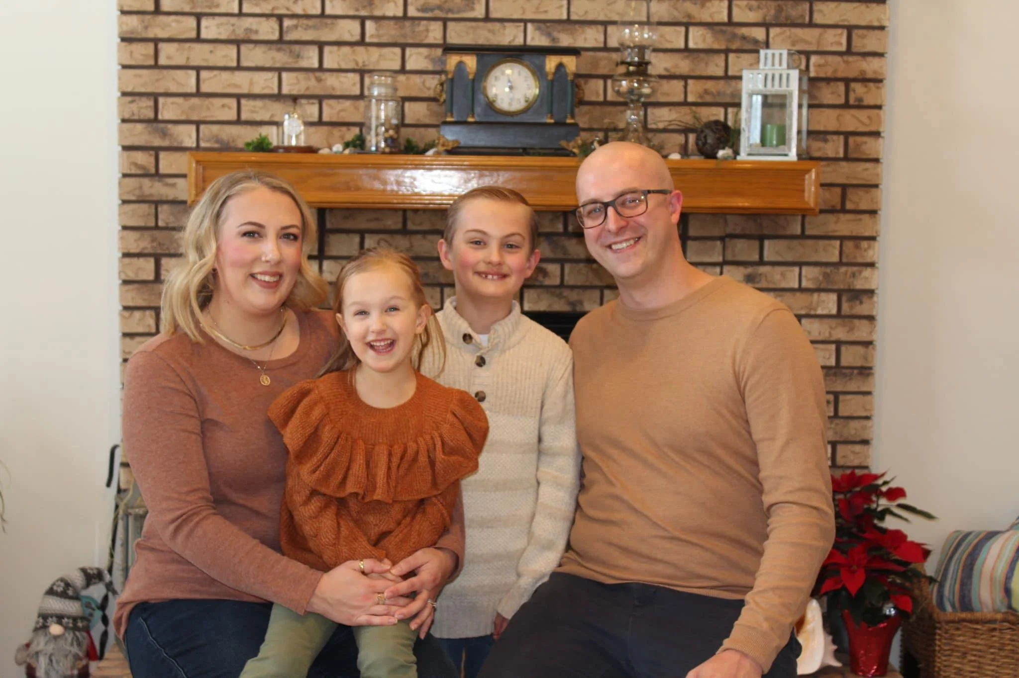 A family of four sitting and standing in front of a brick fireplace, smiling for a photo in a living room decorated with holiday decor, including poinsettias and candles.