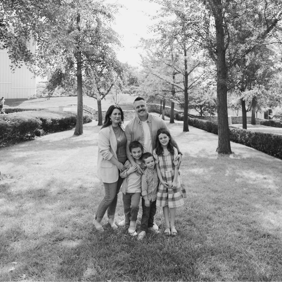 A family of six standing on a grassy park with trees and shrubs in the background, black and white photo, smiling and posing for the camera.
