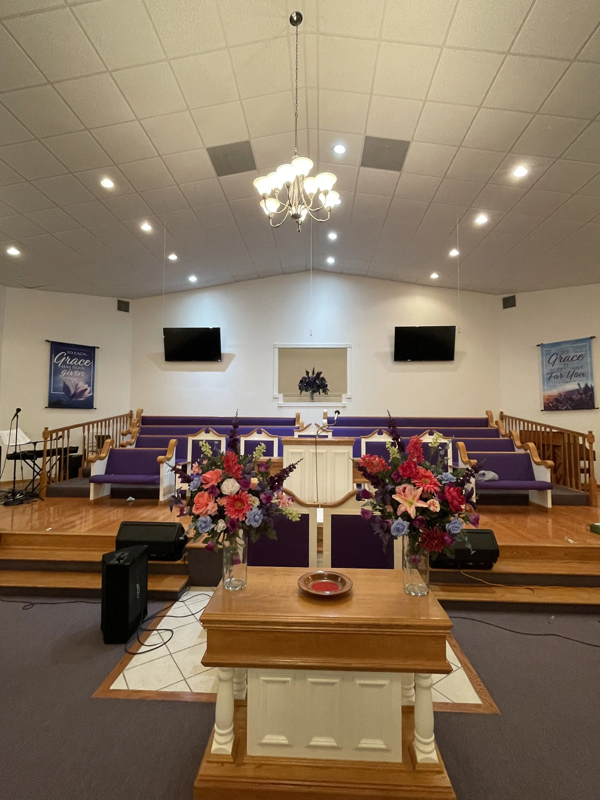 Interior of a church sanctuary with purple pews, a wooden lectern, floral arrangements, and two large screens on the wall.