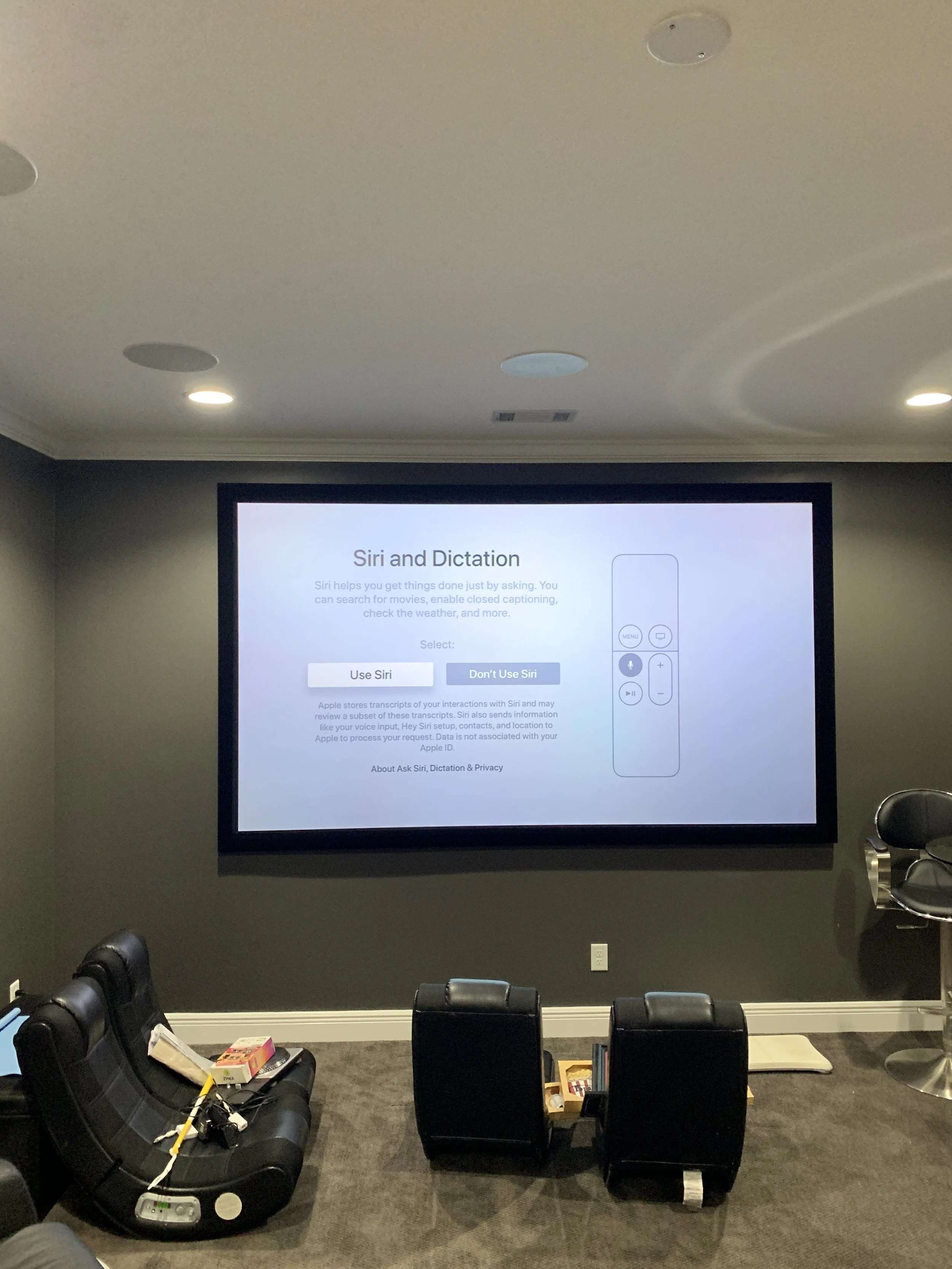 Living room with a large television screen displaying Siri and Dictation settings, surrounded by black chairs, a black couch with items, and a dark gray wall.