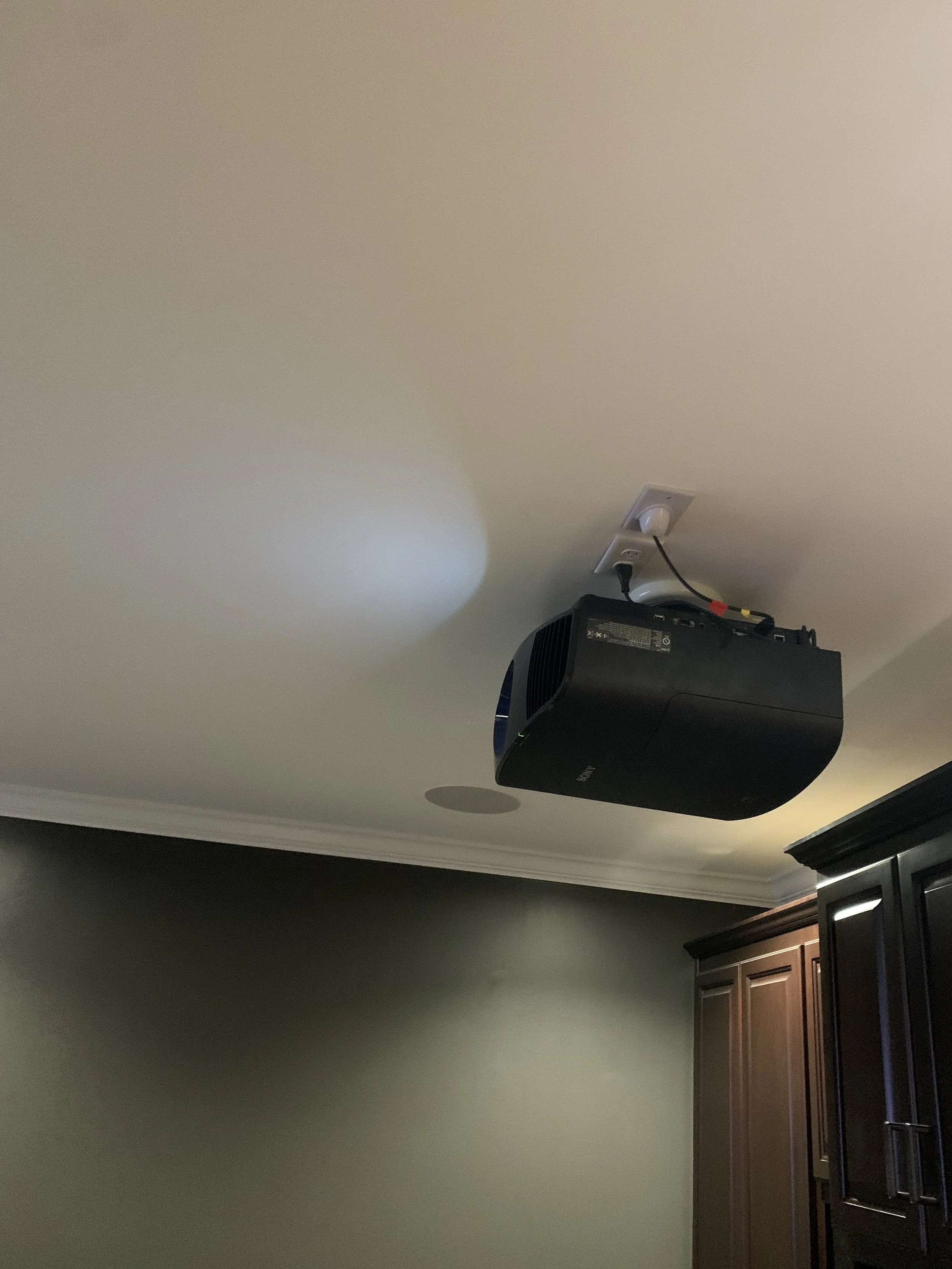 Ceiling-mounted black projector with power and video cables connected, in a room with a beige ceiling, crown molding, and dark brown cabinets.