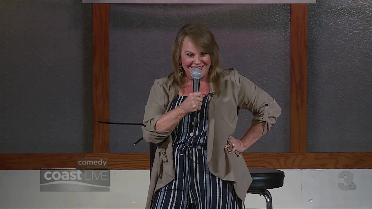 A woman standing on stage, holding a microphone, smiling and wearing a beige jacket over a striped jumpsuit, with her left hand on her hip, in front of a dark gray wall with wooden accents.