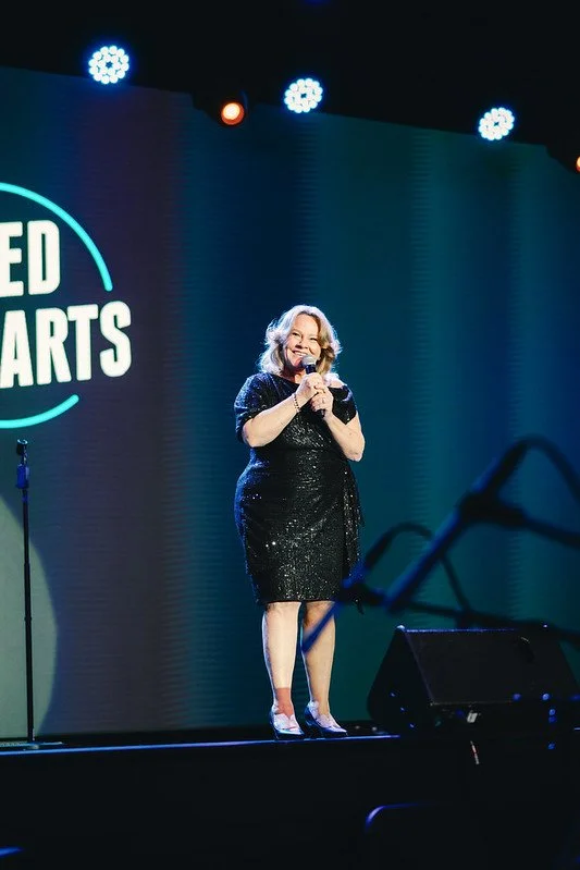 A woman in a black, sequined dress and heels standing on stage, holding a microphone, and smiling. There is a large screen behind her with the words "United Hearts" and stage lights overhead.