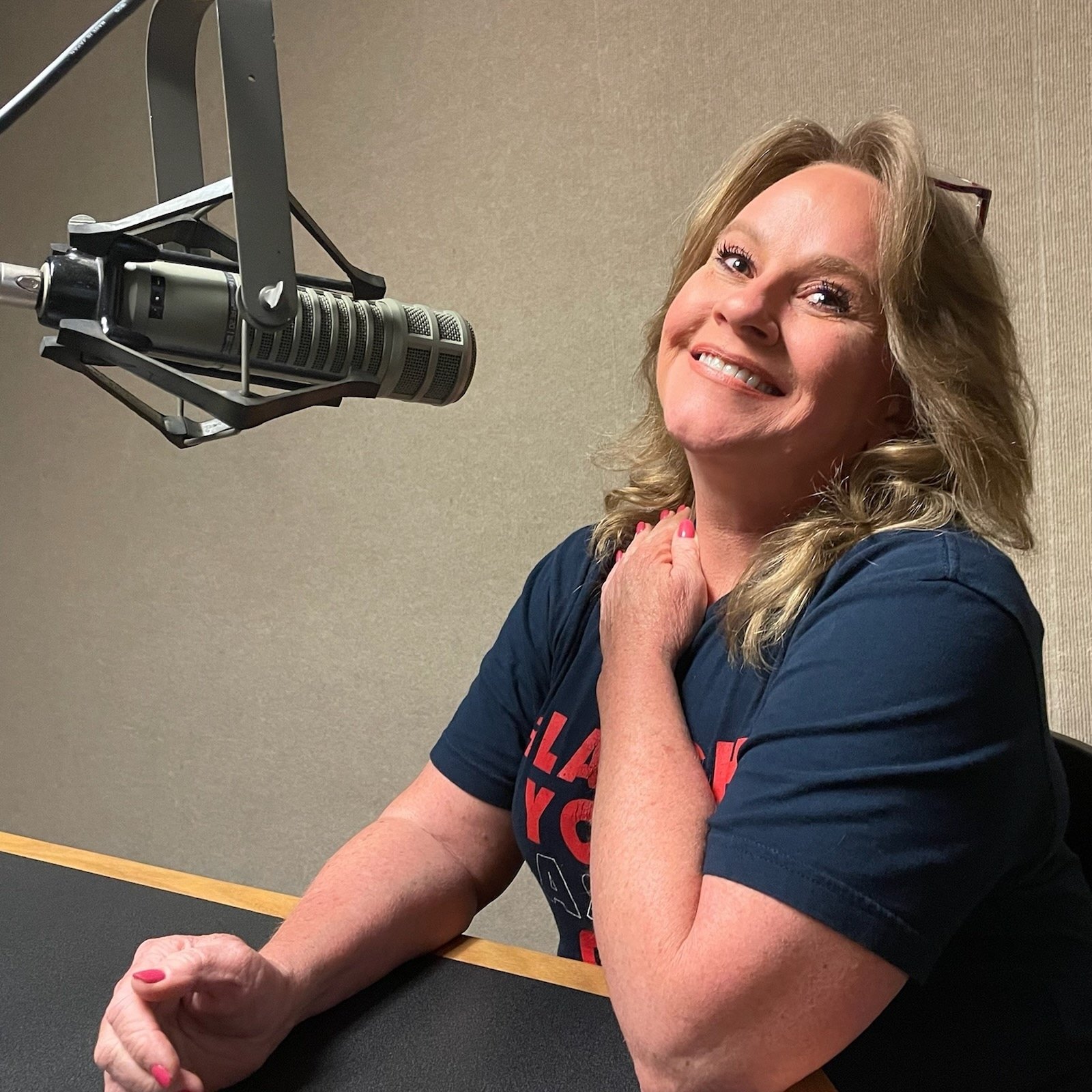 A woman with blonde hair smiling, sitting at a table in front of a microphone in a recording studio or radio station.