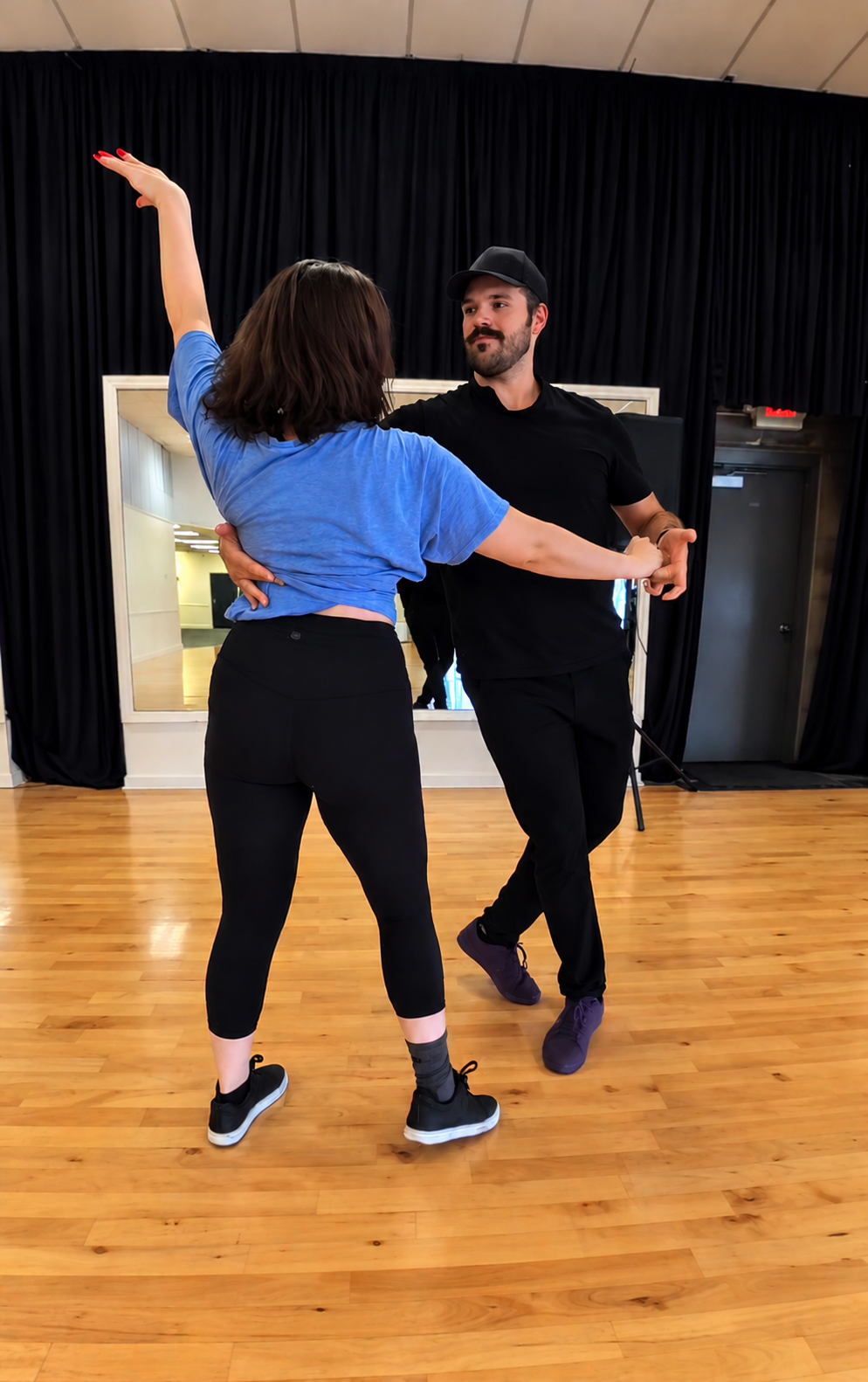 A man and a woman dance together in a dance studio with a wooden floor and a large mirror in the background.