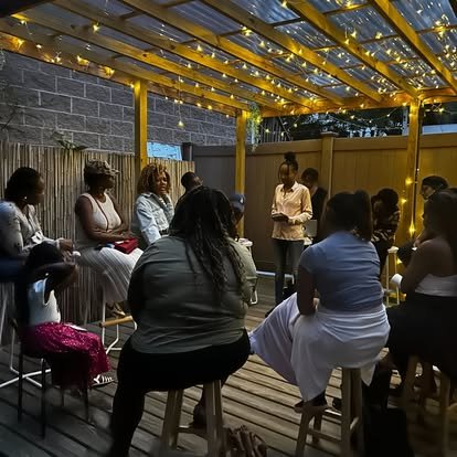 A group of women sitting in a circle under a gazebo decorated with string lights, participating in a gathering or discussion outdoors at night.