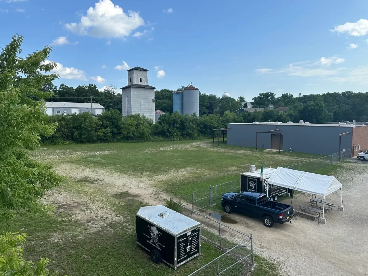 An outdoor scene with a large open grassy and dirt area, a black truck with a white tent, and a food truck with an ice cream image, fenced in with a chain-link fence. Behind are industrial buildings, silos, trees, and a blue sky with scattered clouds