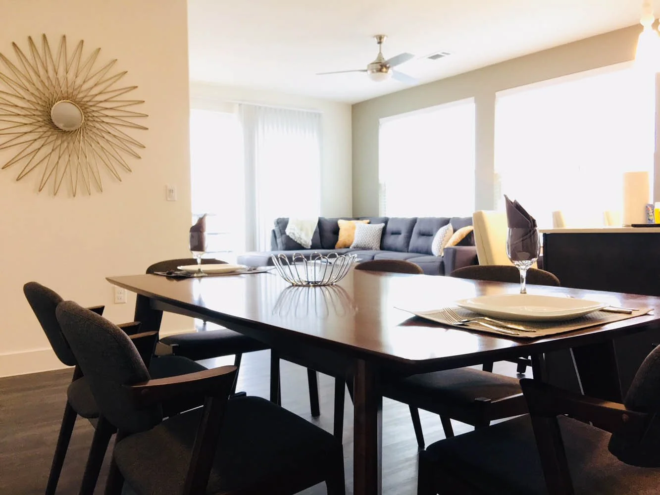 Dining area with a wooden table set with plates, silverware, and wine glasses, surrounded by chairs, and a living room with a gray sectional sofa, available to rent at Furniture Options.